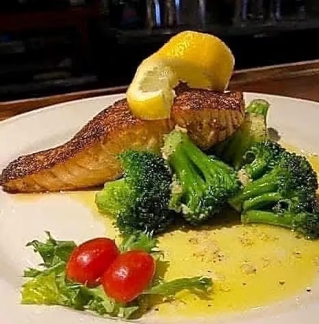 Grilled fish with lemon wedge, broccoli, cherry tomatoes, and sauce on a white plate.
