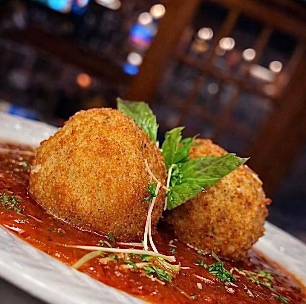 Three fried croquettes garnished with fresh basil leaves on a plate with sauce, in a dimly lit restaurant setting.