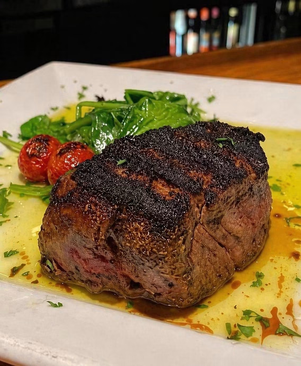 Cooked steak with char marks, served with greens and cherry tomatoes on a white plate.
