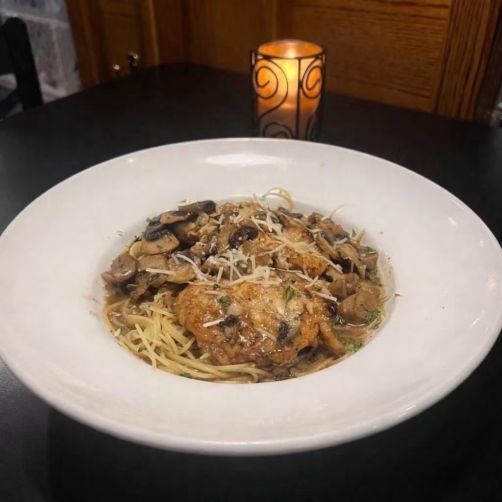 Plate of pasta with sauce, mushrooms, and grated cheese on a dark table, candle in background, warm lighting.