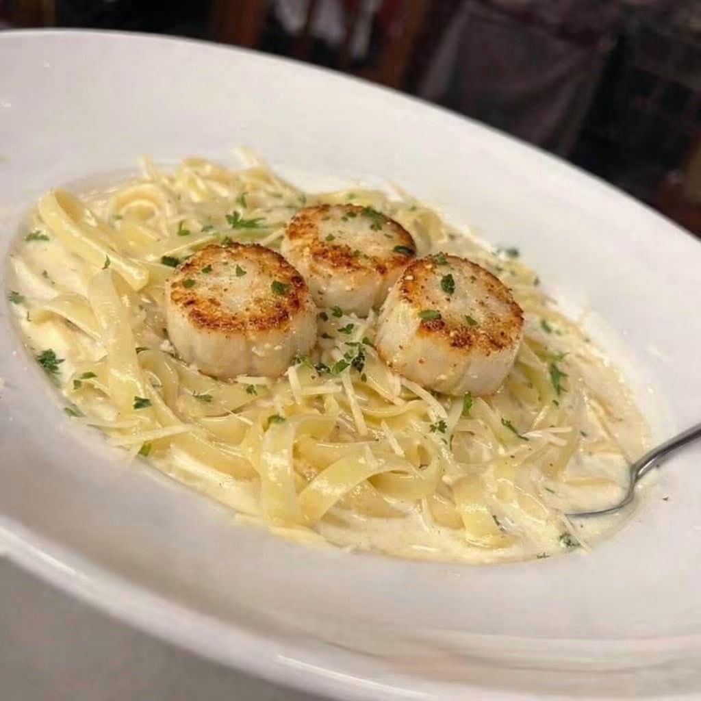 Fettuccine pasta with three seared scallops on top, served in a white bowl, garnished with herbs.
