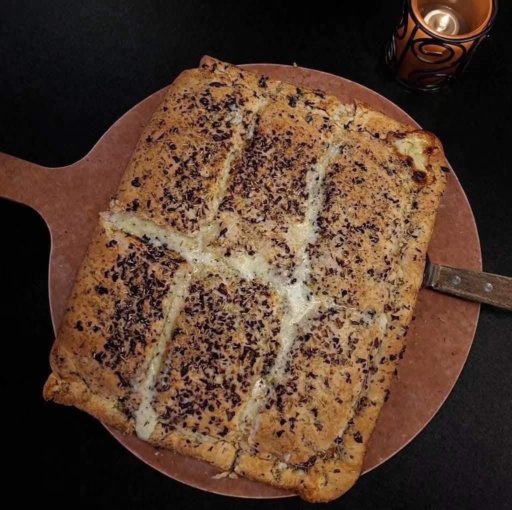 Large baked dish with a golden crust, topped with chocolate sprinkles, on a round wooden board with a knife, next to a drink.