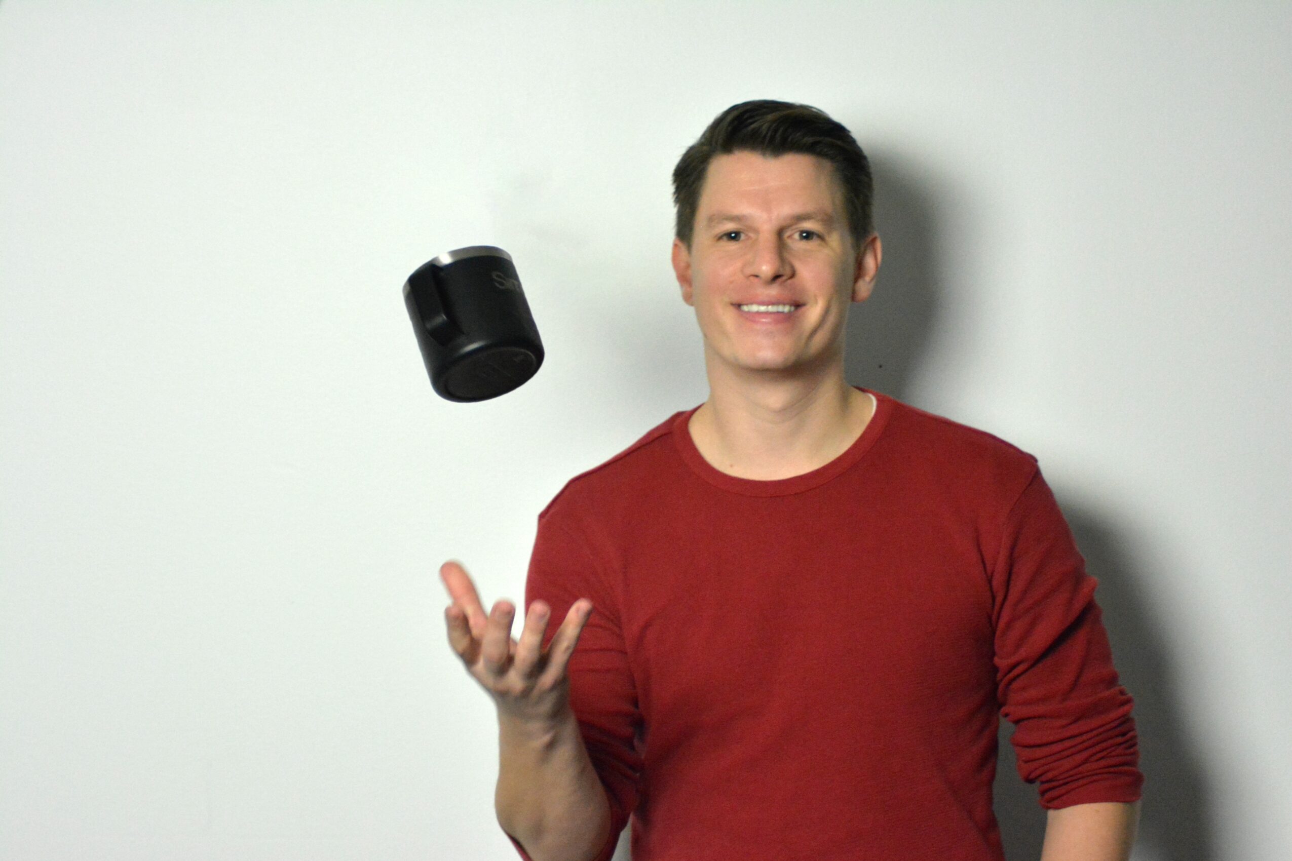 Man in red shirt tossing a black camera in the air, smiling, against a plain light background.