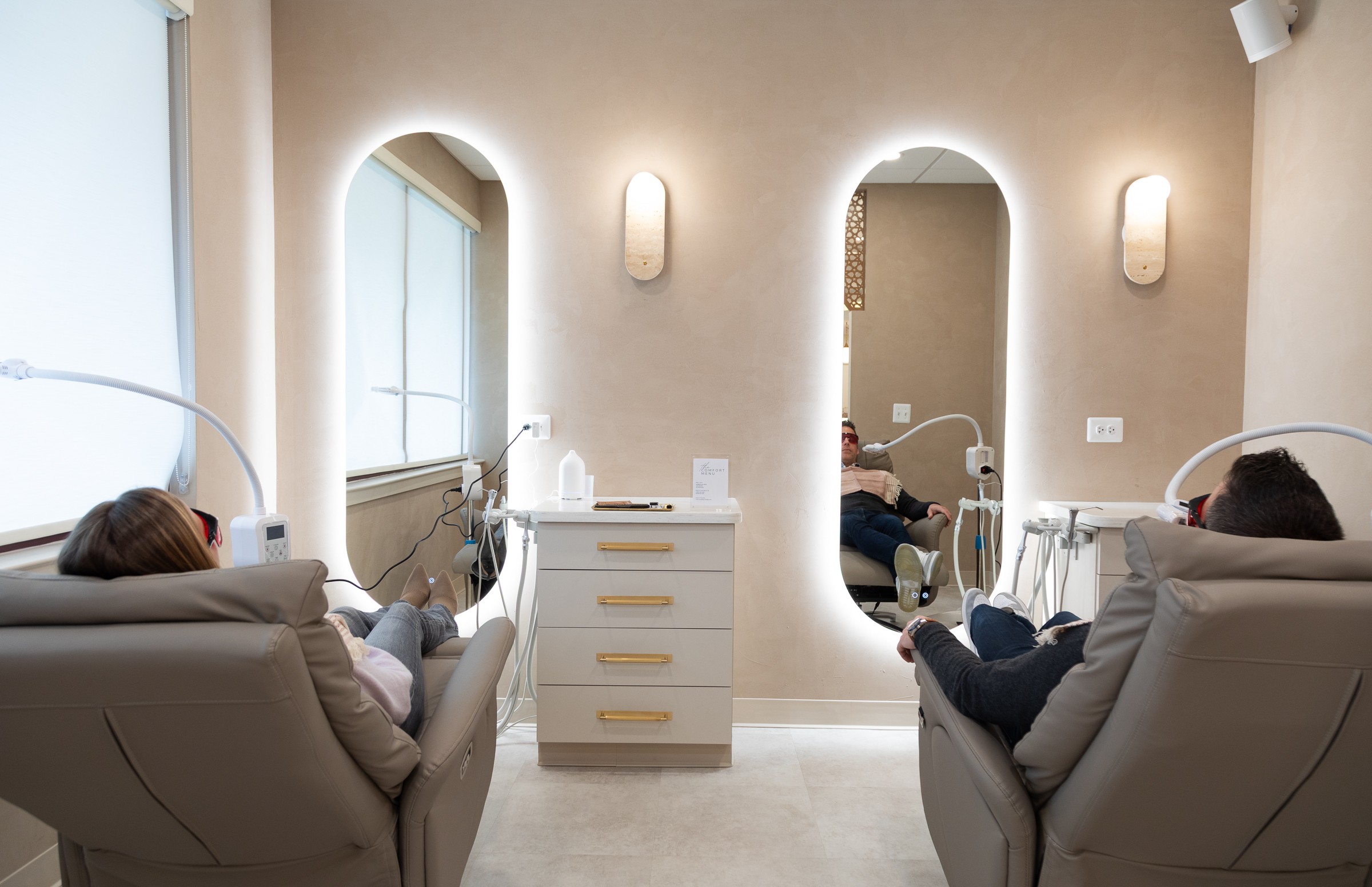Two people receiving treatment in reclining chairs in a clean, well-lit room with arched mirrors and wall-mounted lights.