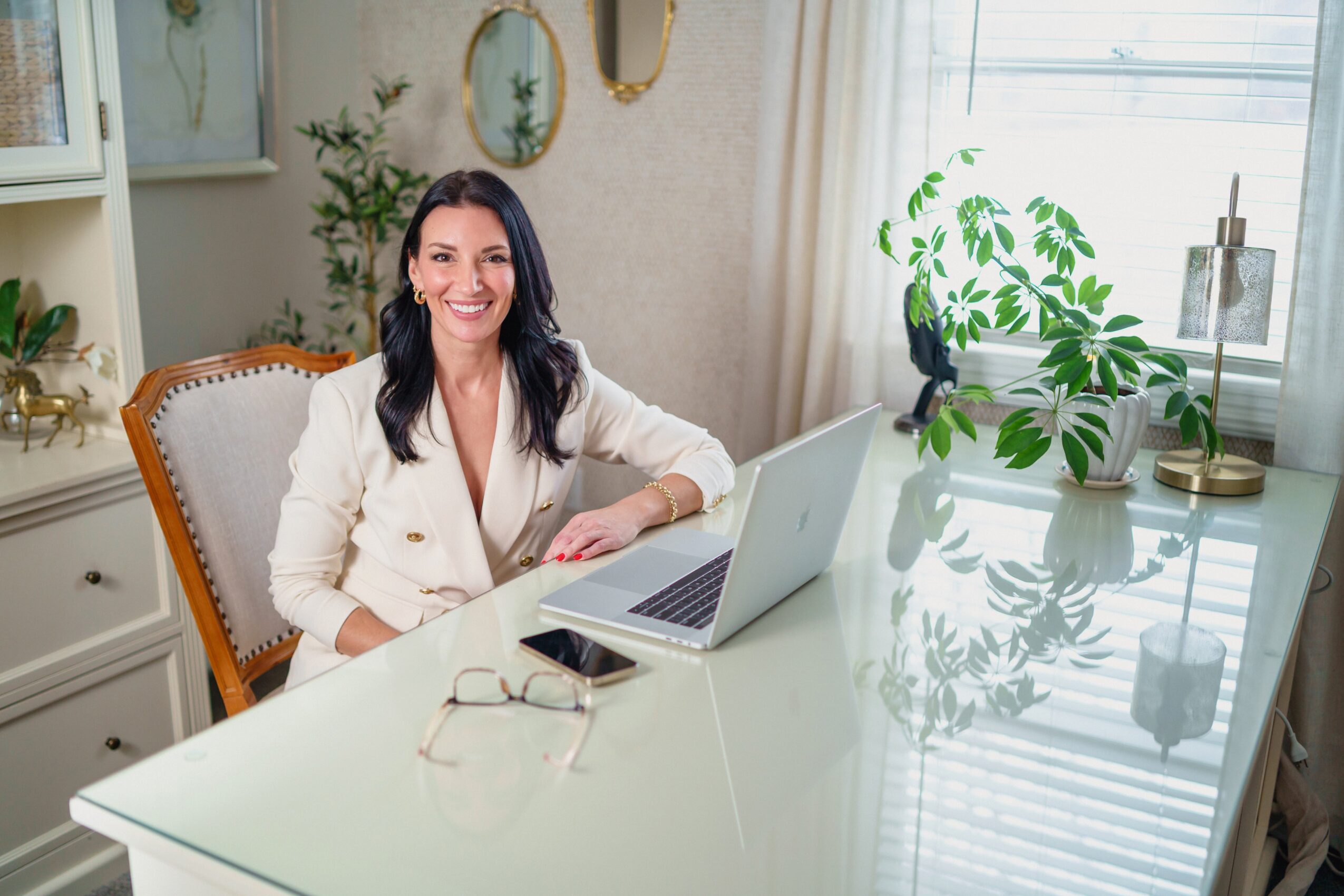 Woman sitting at a desk with a laptop, smiling, in a bright room with plants and decor, sunlight streaming in.