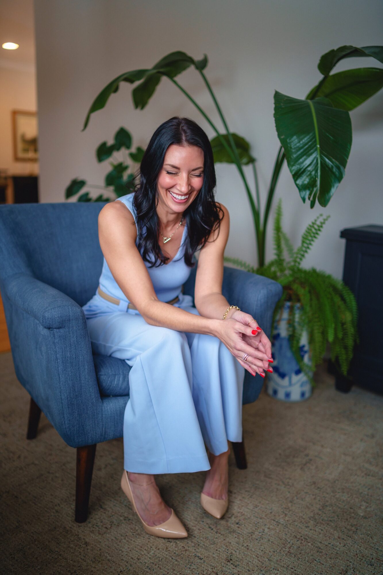 Woman sitting on a blue armchair, smiling, with large green plants behind her, wearing a light blue outfit and beige heels.