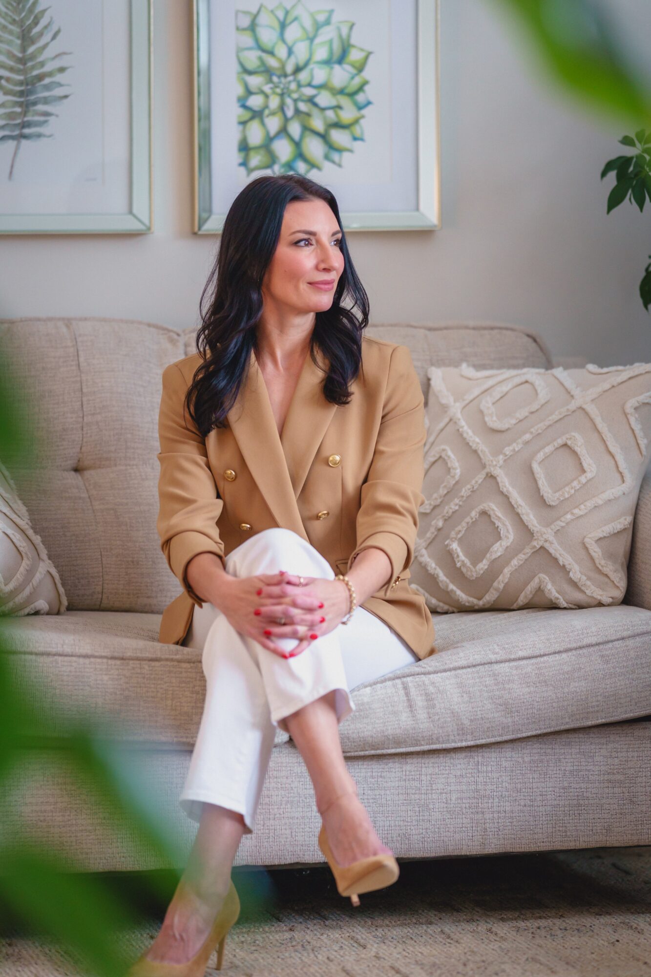 Woman sitting on sofa holding a mug, with framed botanical art behind her, beige and white decor, and green plants nearby.