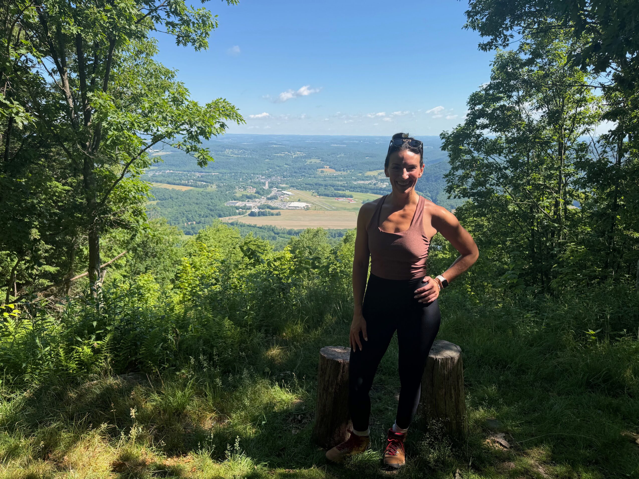Woman standing outdoors on a tree stump with a landscape view, trees, and blue sky in the background.