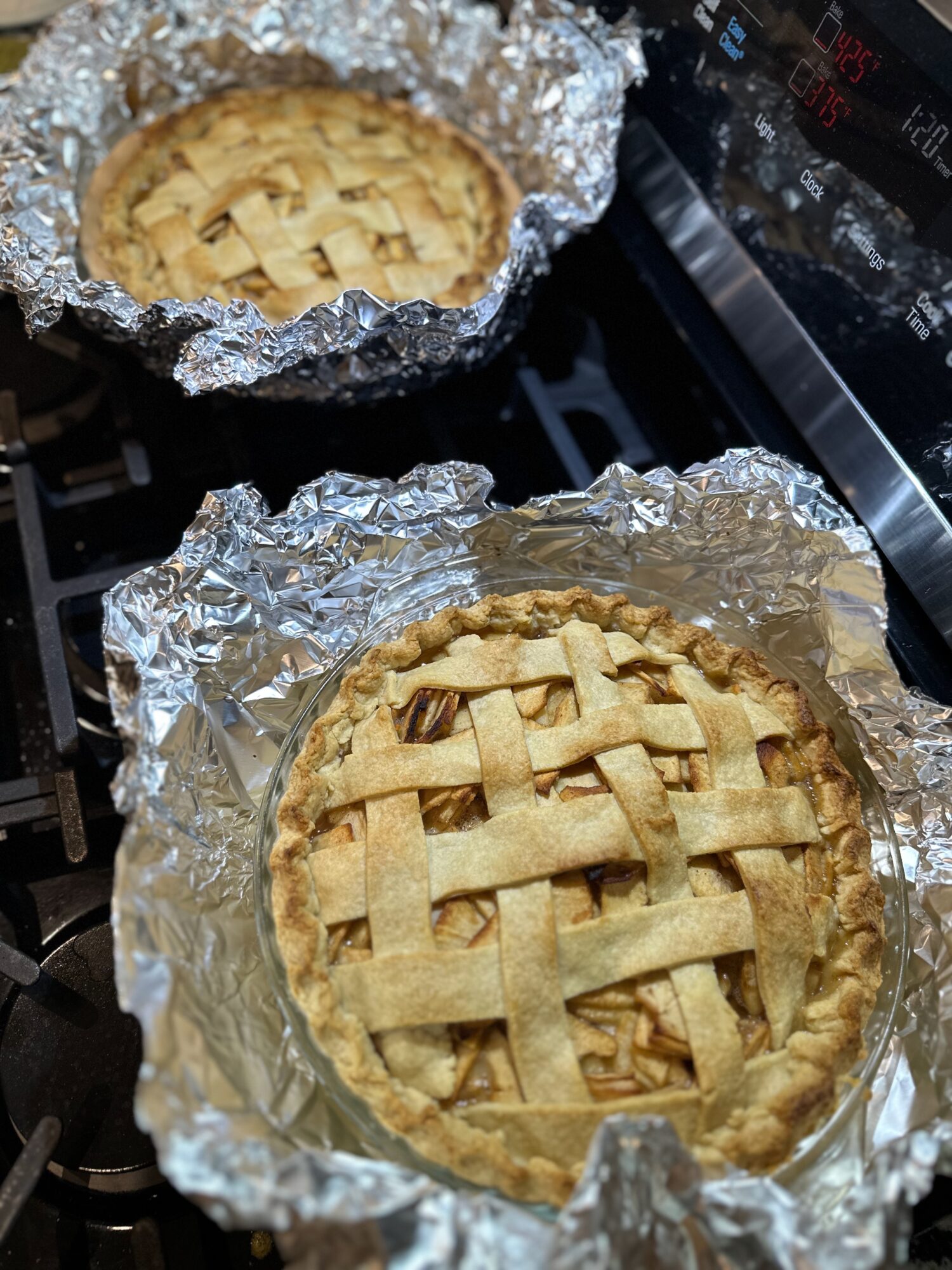 Two mini pies in aluminum foil on a stovetop, with lattice crusts visible, one in the foreground and one in the background.
