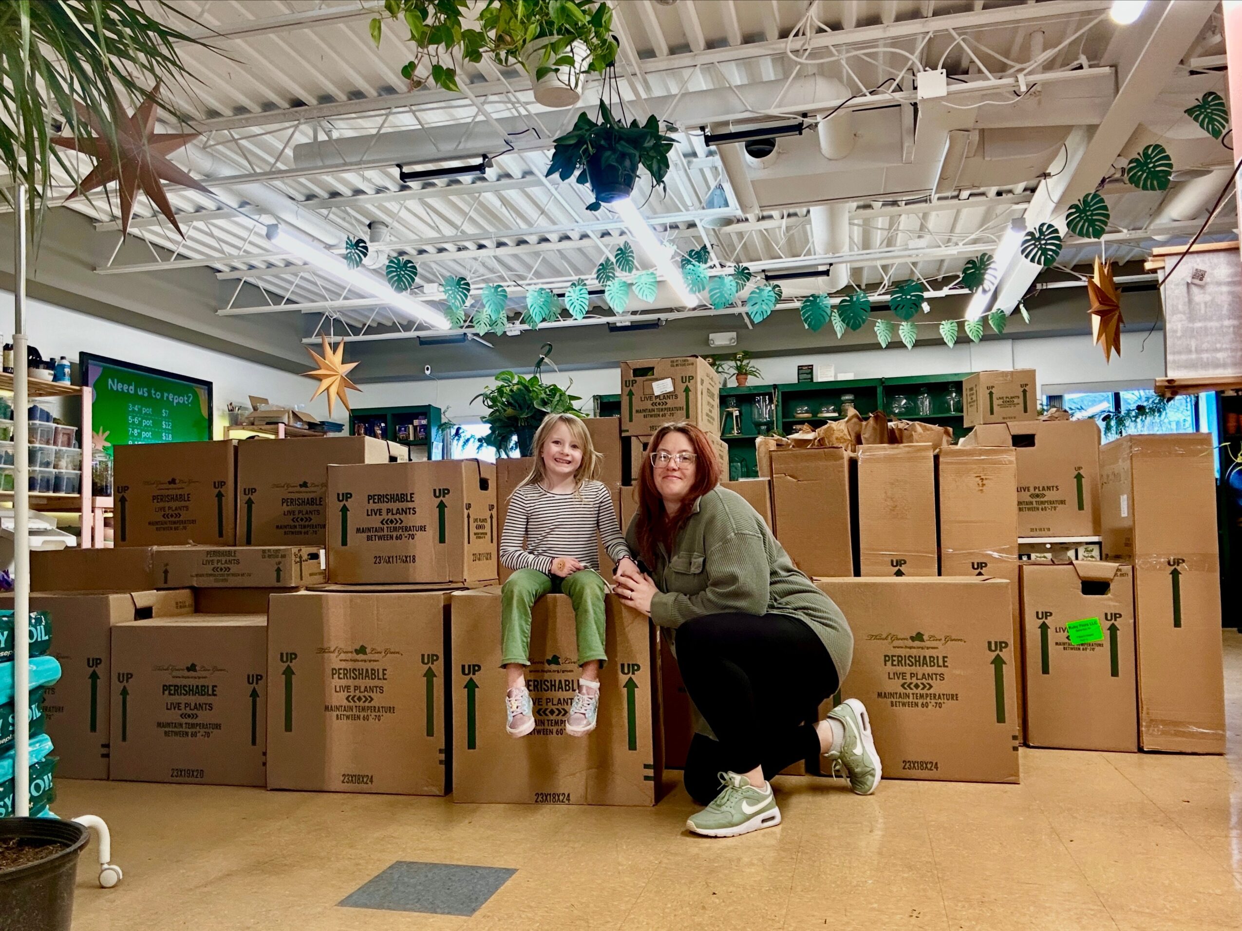 Two women sit on boxes in a store decorated with hanging paper and star decorations, surrounded by stacked boxes.