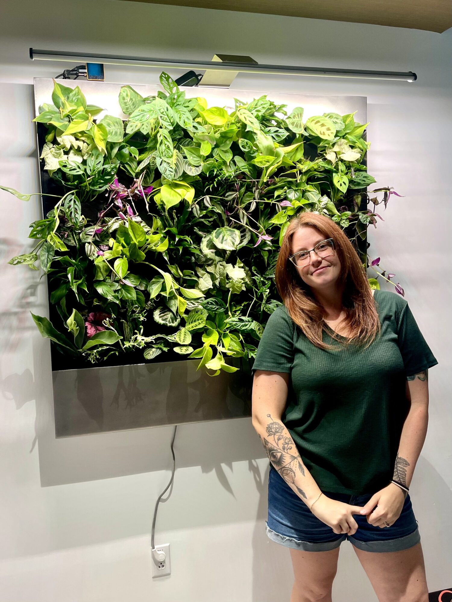 Woman with glasses and tattoos standing in front of a vertical garden with green plants, smiling.
