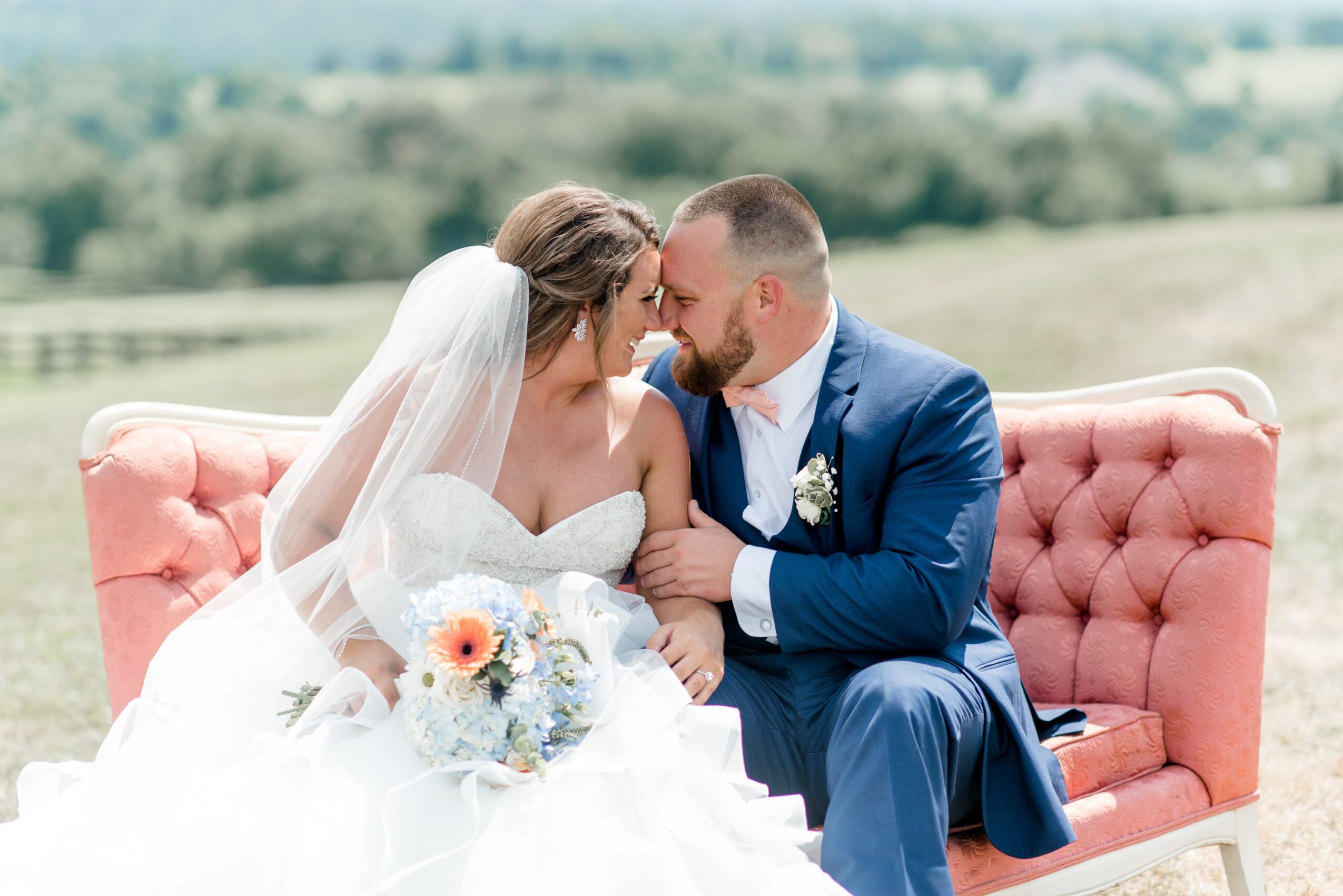 Bride and groom sitting on a pink sofa outdoors, smiling and leaning close, with a scenic landscape in the background.