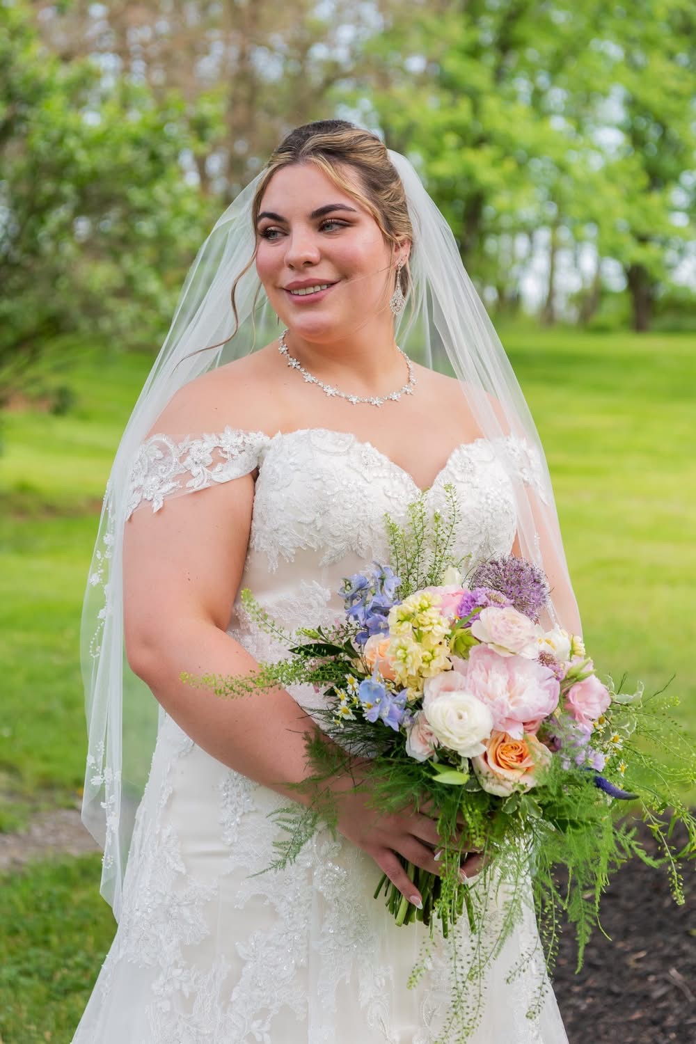 Bride in wedding dress holding a bouquet outdoors with trees and grass in background.