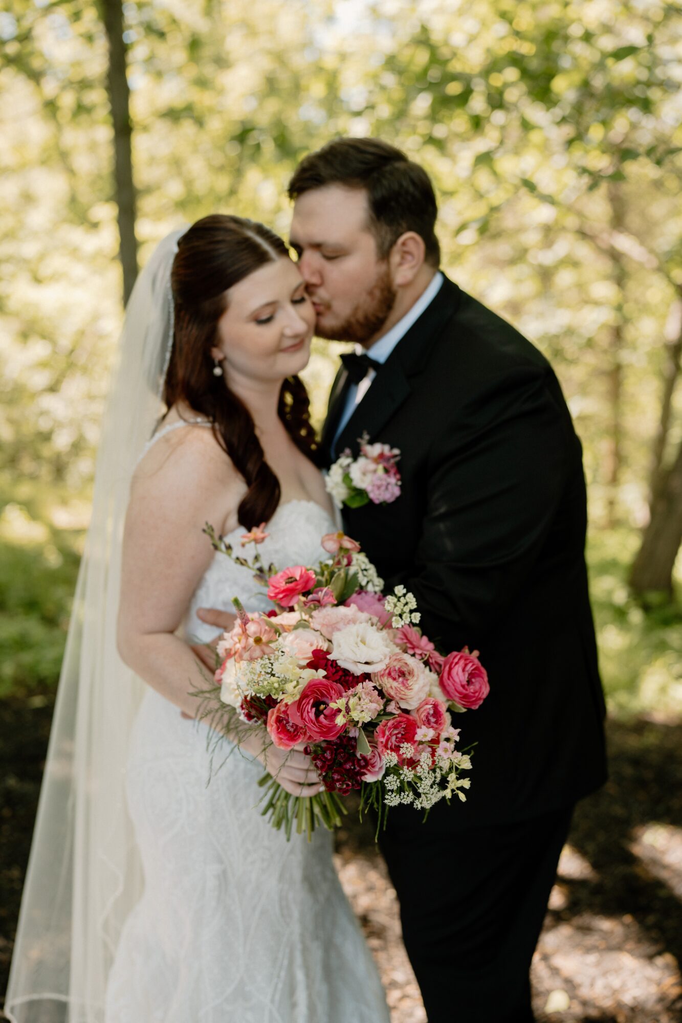 A bride and groom in a forest, the groom leaning close to the bride holding a large bouquet of pink and white flowers.