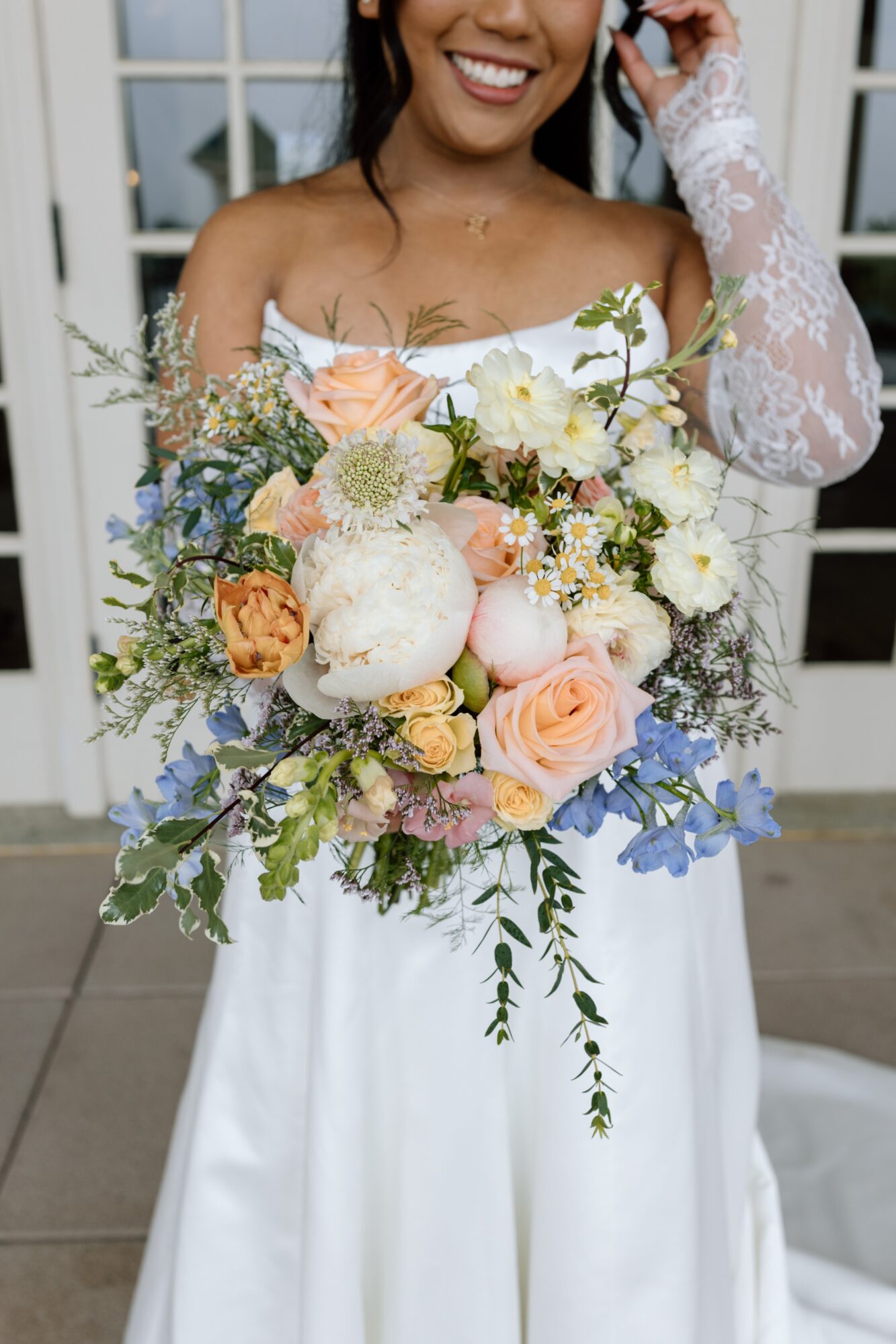 Woman in wedding dress holding a large bouquet of flowers, smiling, with a building in the background.