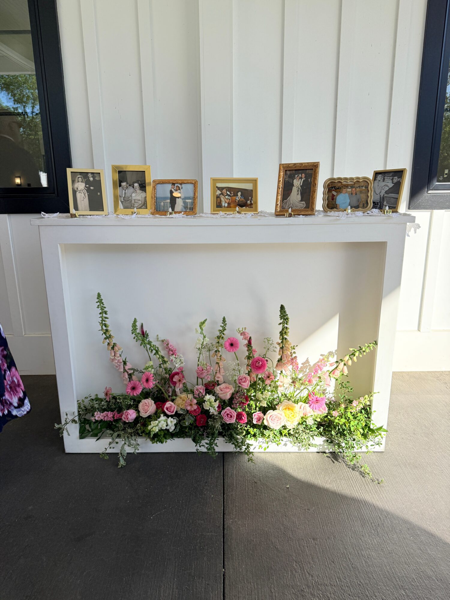 White display shelf with framed photos and a floral arrangement of pink, white, and green flowers, outdoors on concrete.