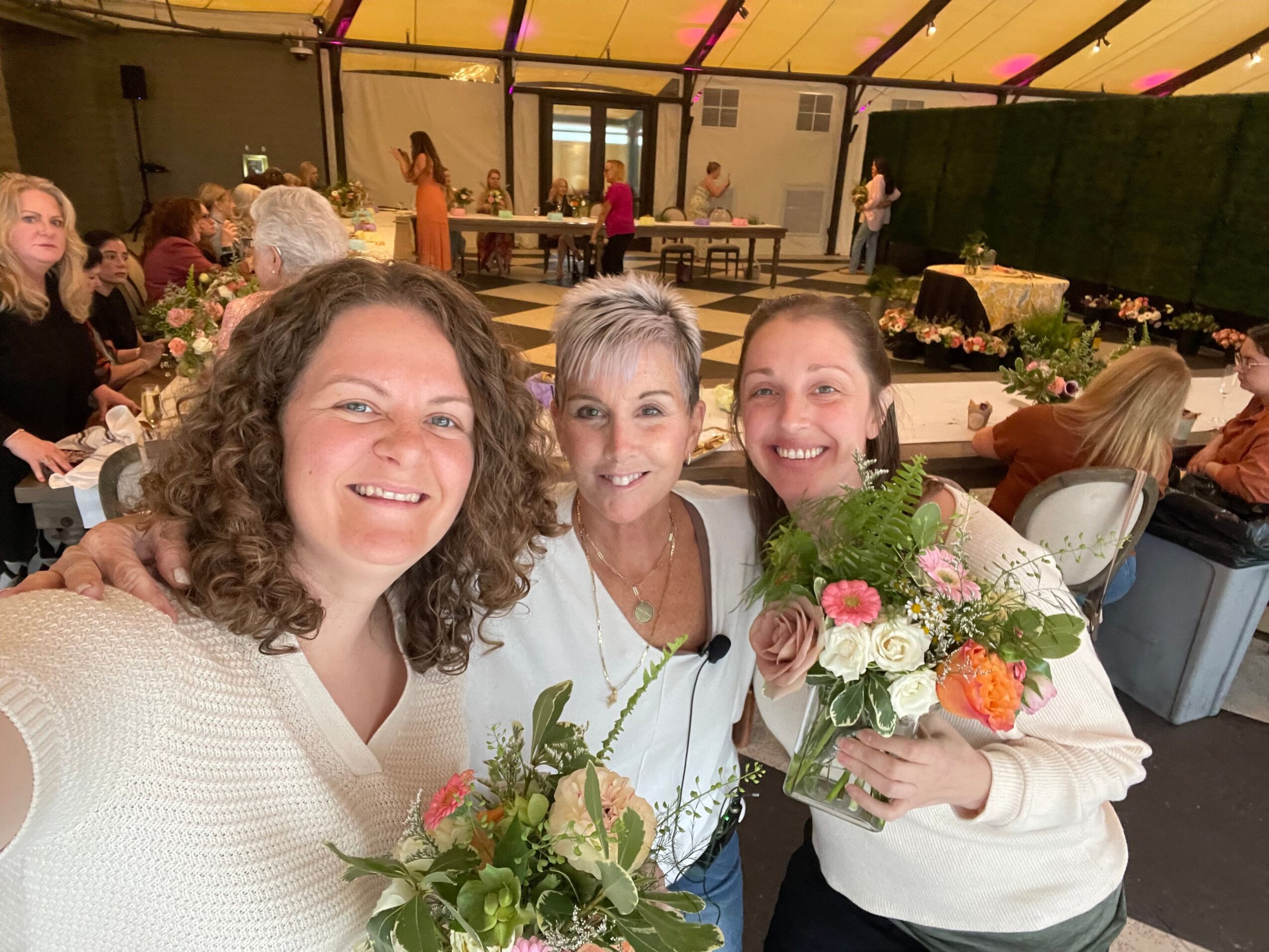 Three women smiling, holding flowers, and standing indoors with a decorated background.