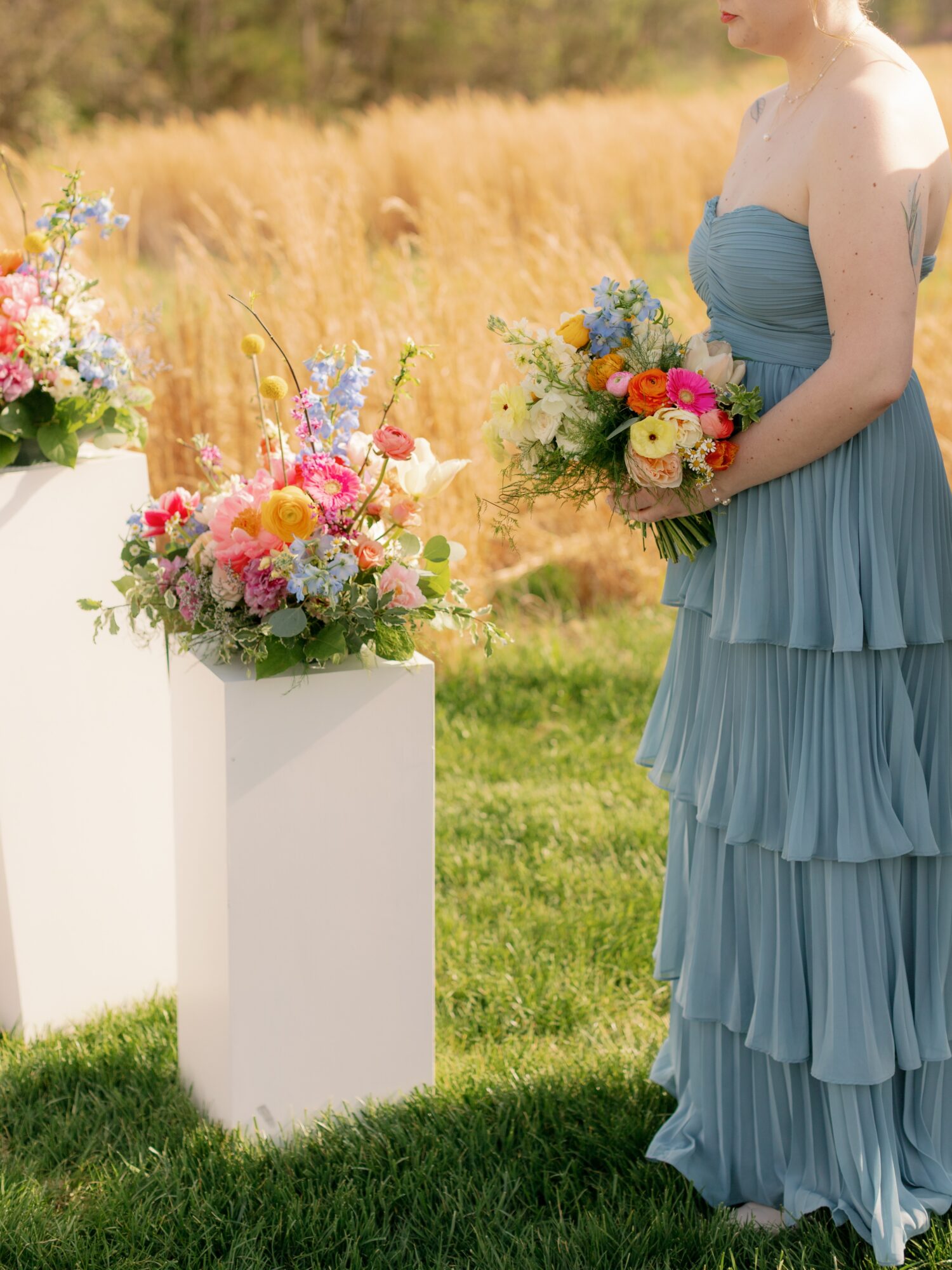 Woman in a blue dress holding a bouquet, standing outdoors near floral arrangements on white pedestals, grassy field background.