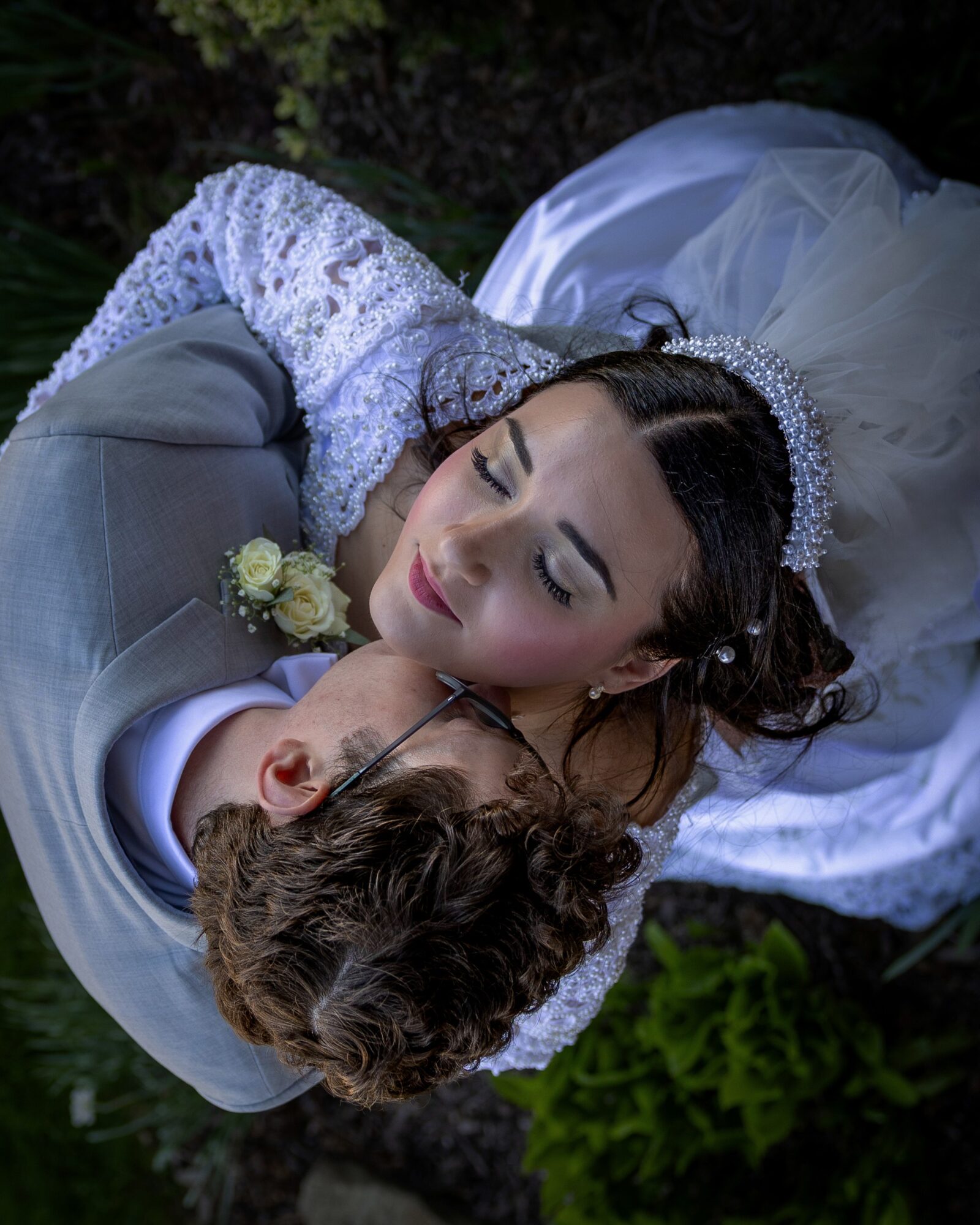 Bride and groom lying outdoors, embracing, with greenery in background, bride wearing a tiara and wedding dress, groom in suit.