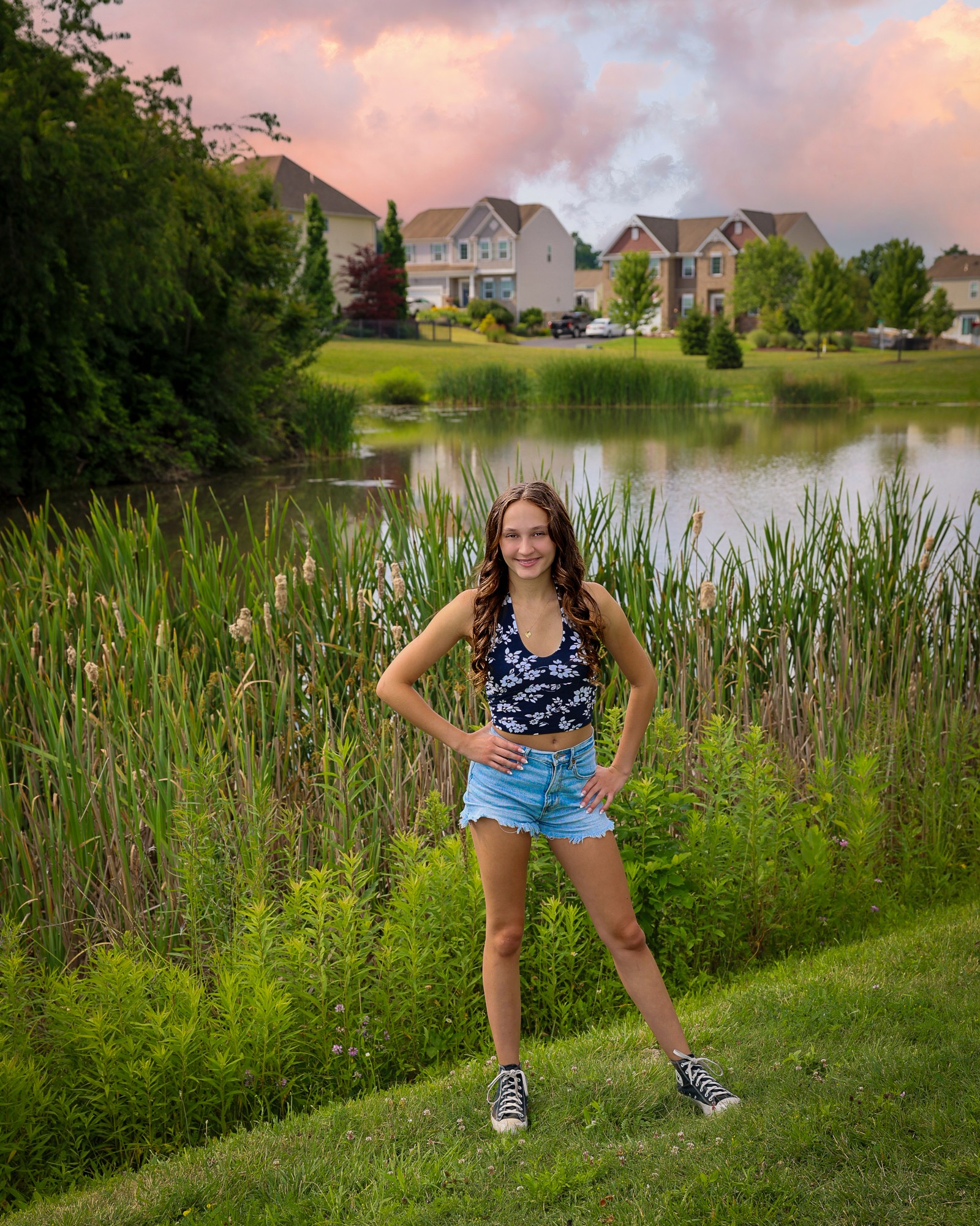 Girl standing on grass near a pond with houses and trees in the background, smiling, hands on hips, wearing a blue top and shorts.