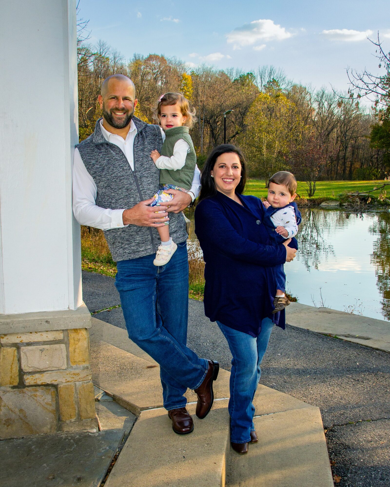 Family of four outdoors near a pond, smiling, with trees and sky in background.