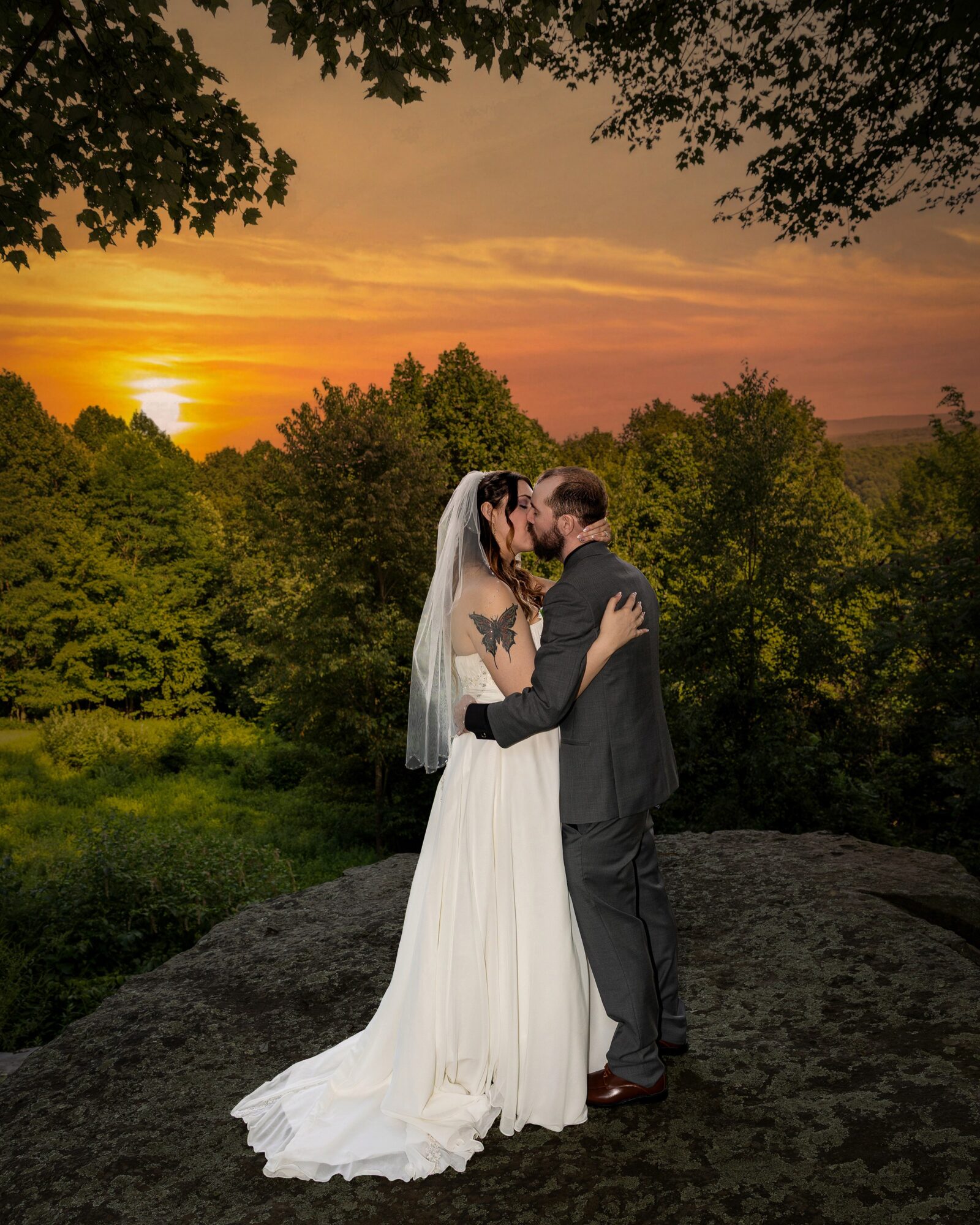 A bride and groom kiss outdoors during sunset, surrounded by trees and greenery, with a cloudy sky in the background.
