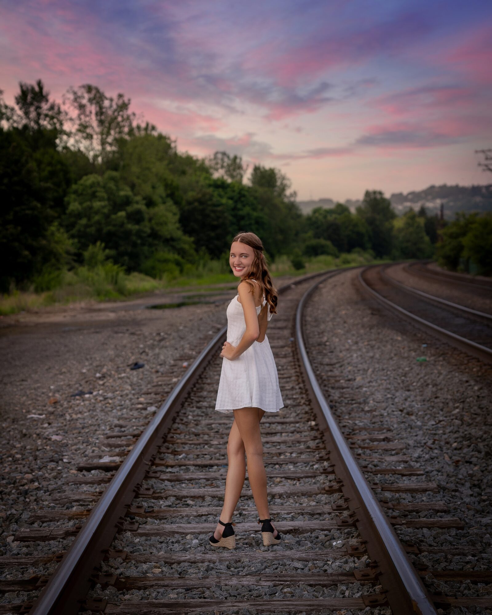 Young woman in a white dress standing on train tracks outdoors during sunset, smiling, with trees and colorful sky in background.