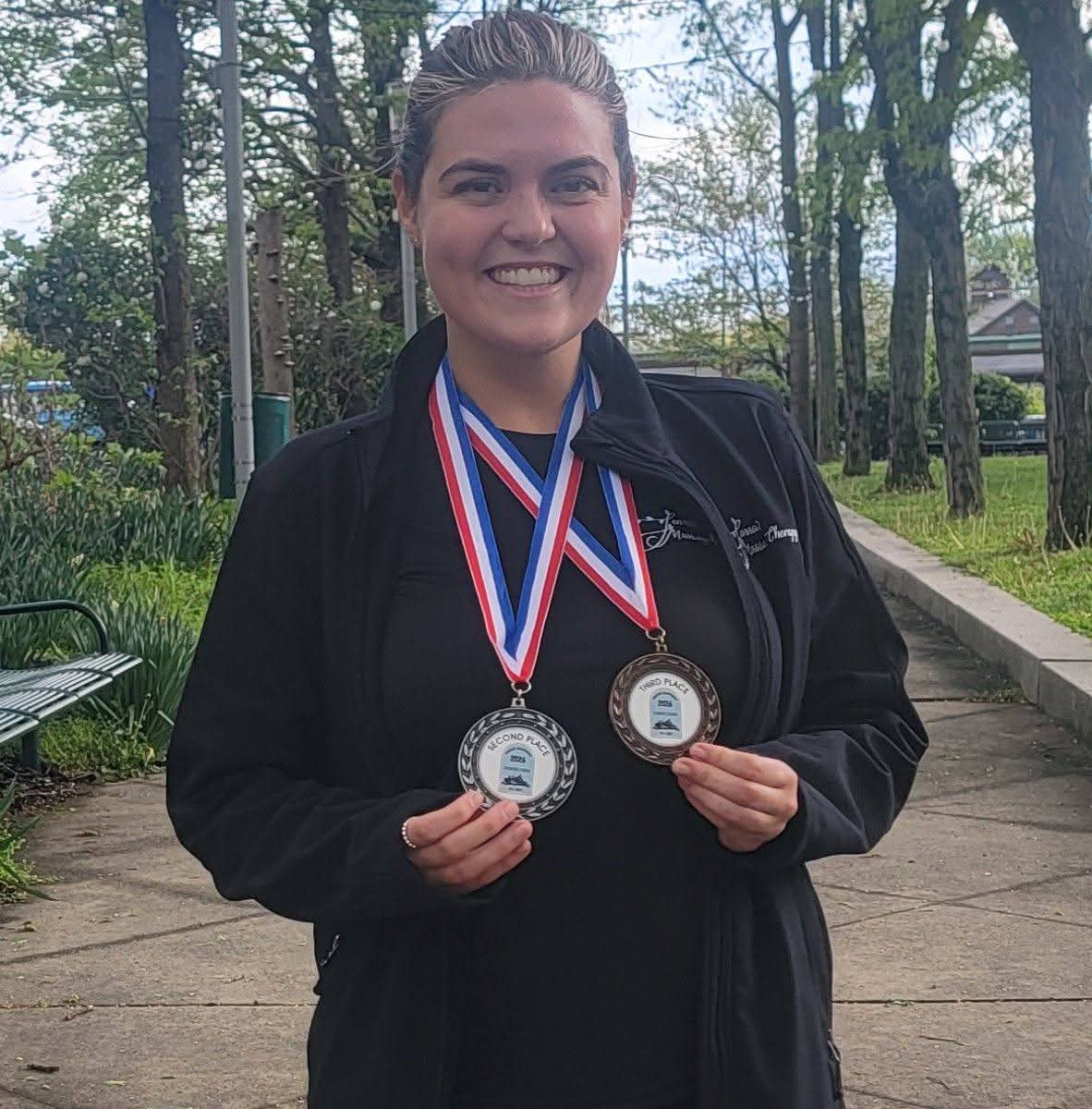 Smiling woman outdoors holding two medals, wearing a black jacket and a black shirt, trees and a pathway in background.