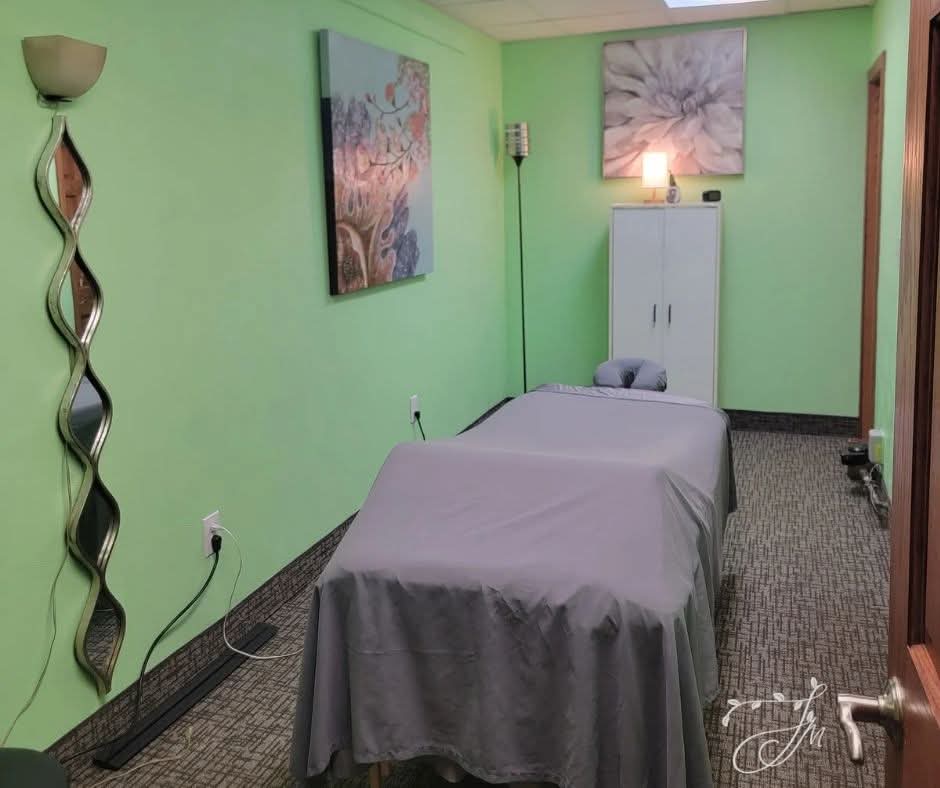 Massage table covered with gray sheet in a room with green walls, artwork, and a cabinet, with a lamp on top.