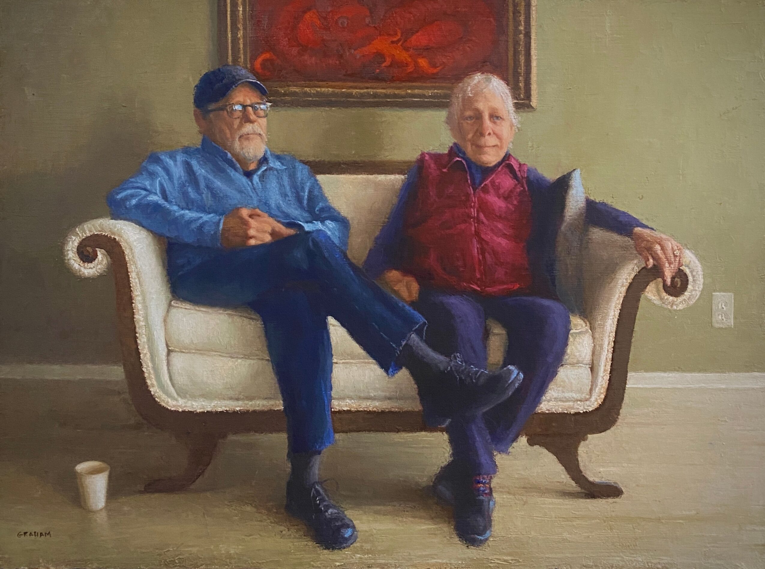Two men sit on a white sofa in a room with green walls and a painting above. One man wears a blue shirt and glasses, the other a red vest. They are smiling.