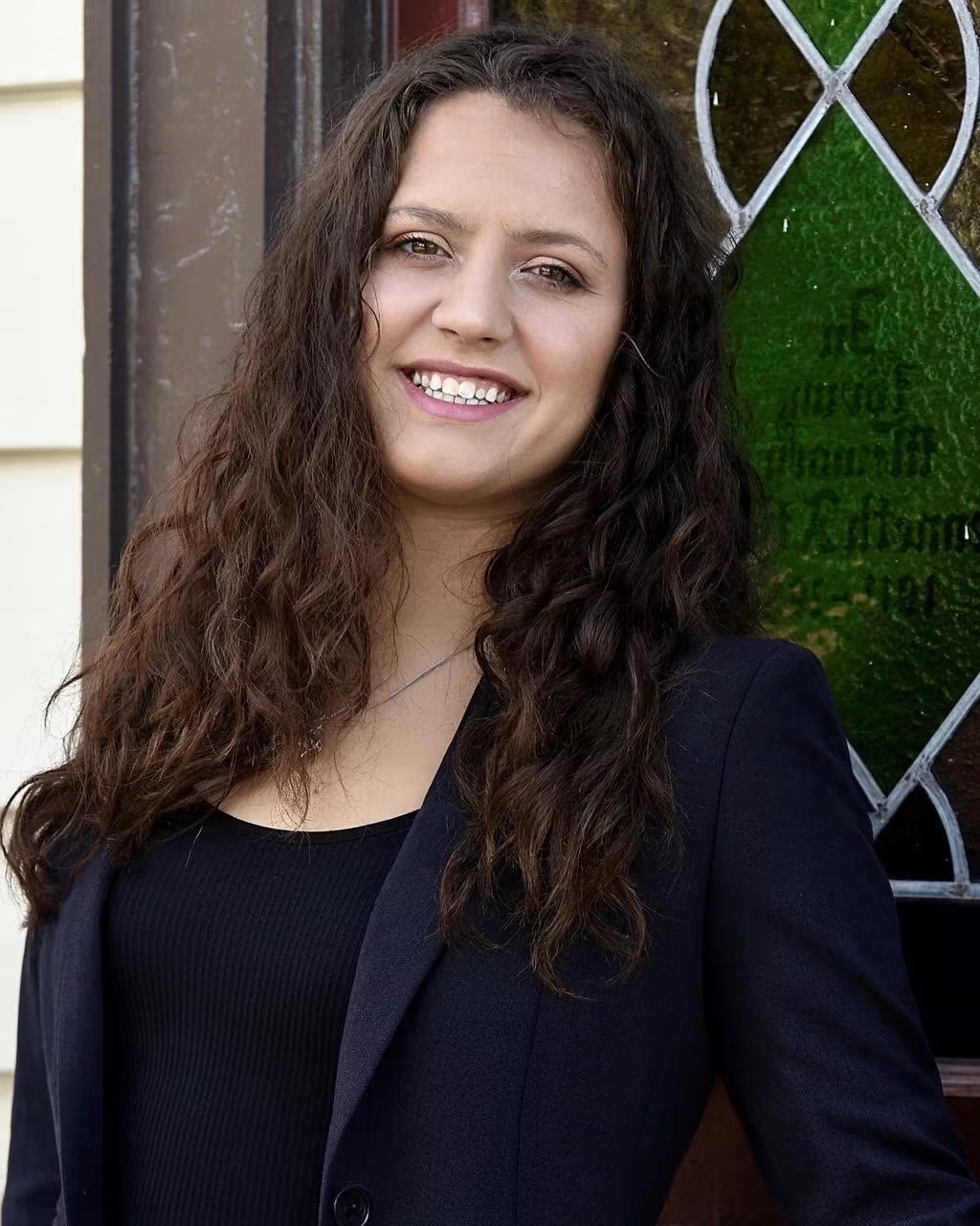 Young woman with long, curly hair smiling outdoors, wearing a dark blazer and top, standing near a decorative window.
