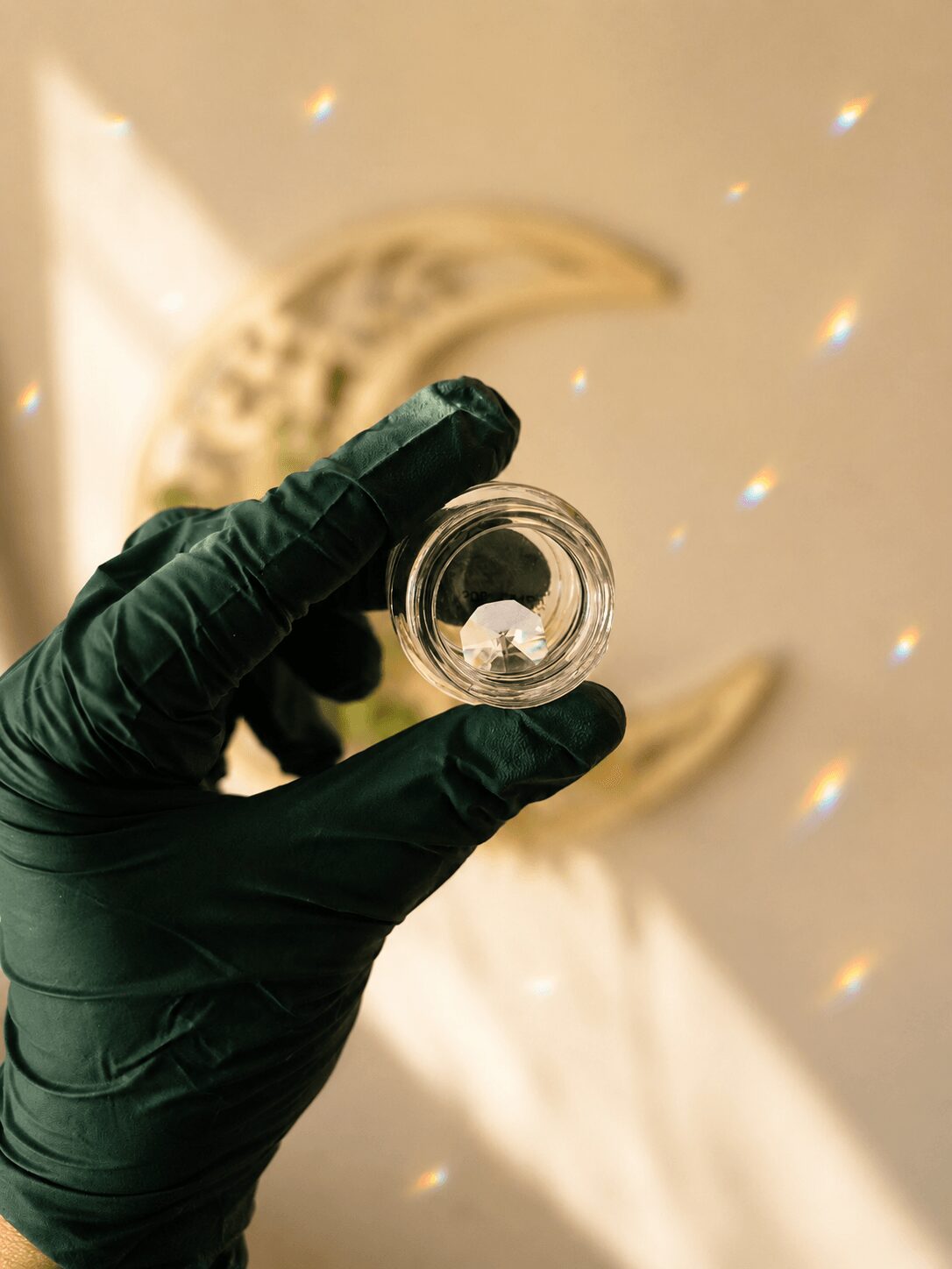 Person wearing a black glove holds a small glass container with a white object inside, against a background with a crescent moon and light reflections.