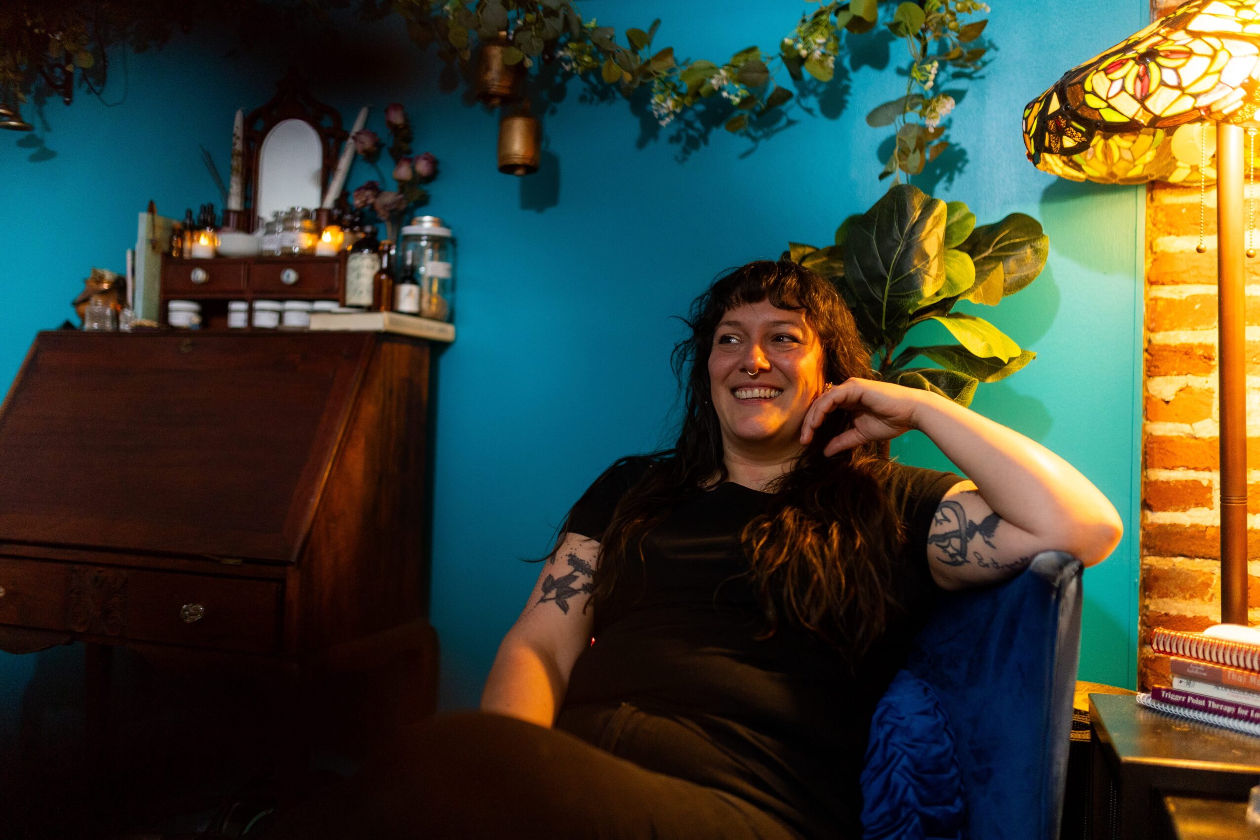 Woman with long dark hair smiling, sitting in a room with blue walls, plants, and warm lighting.