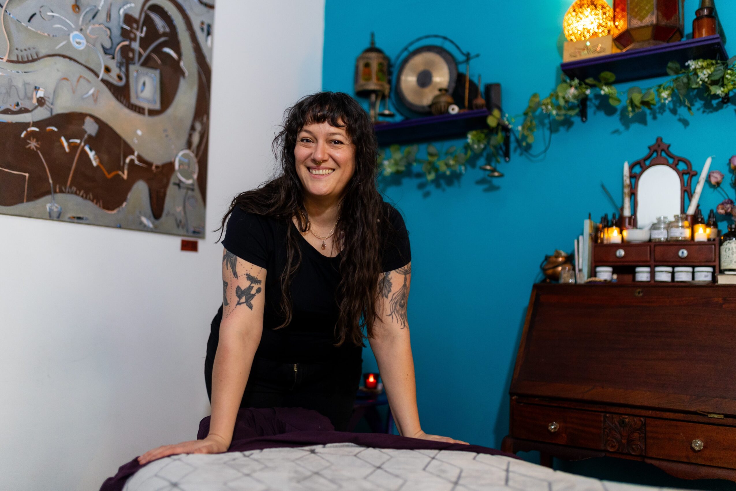 Woman with dark hair and tattoos smiling in a colorful room with blue walls, artwork, and decorative items.