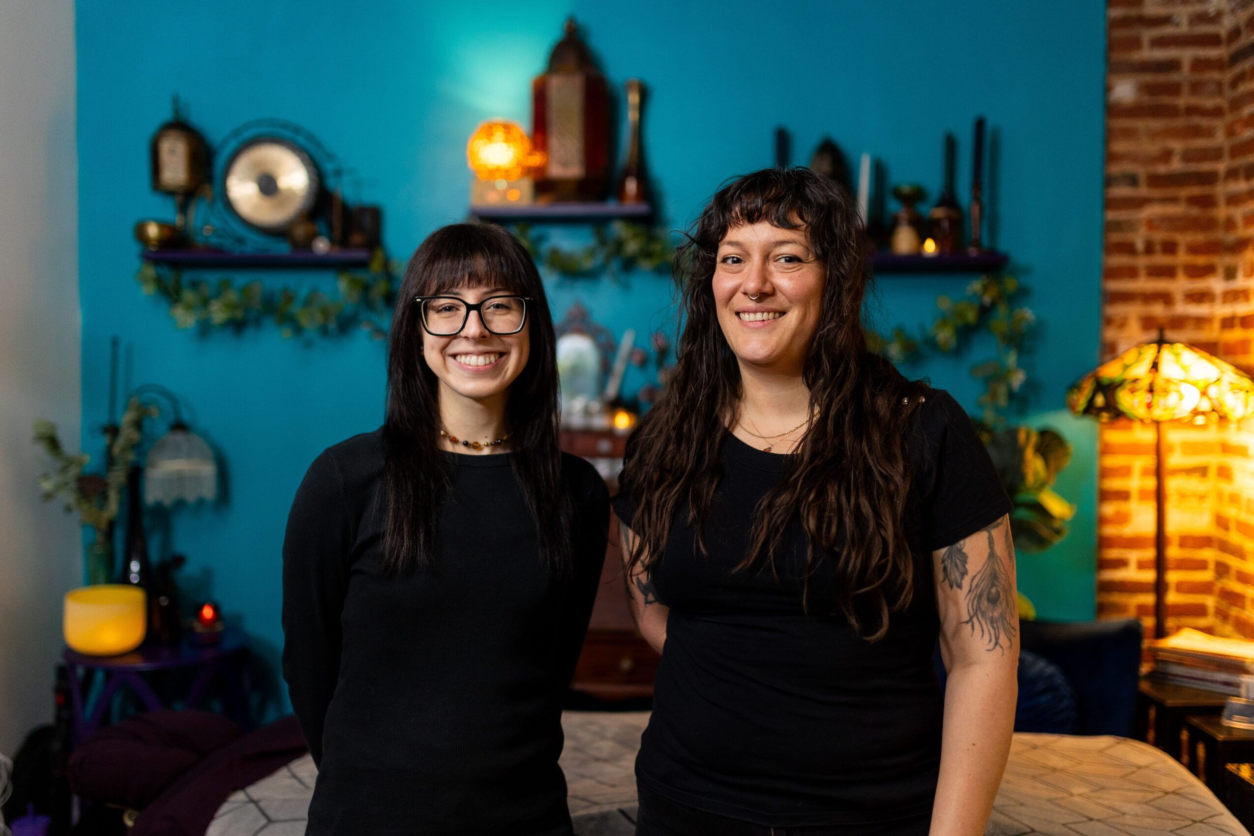 Two women with dark hair and glasses smiling indoors, background with shelves, plants, and warm lighting.