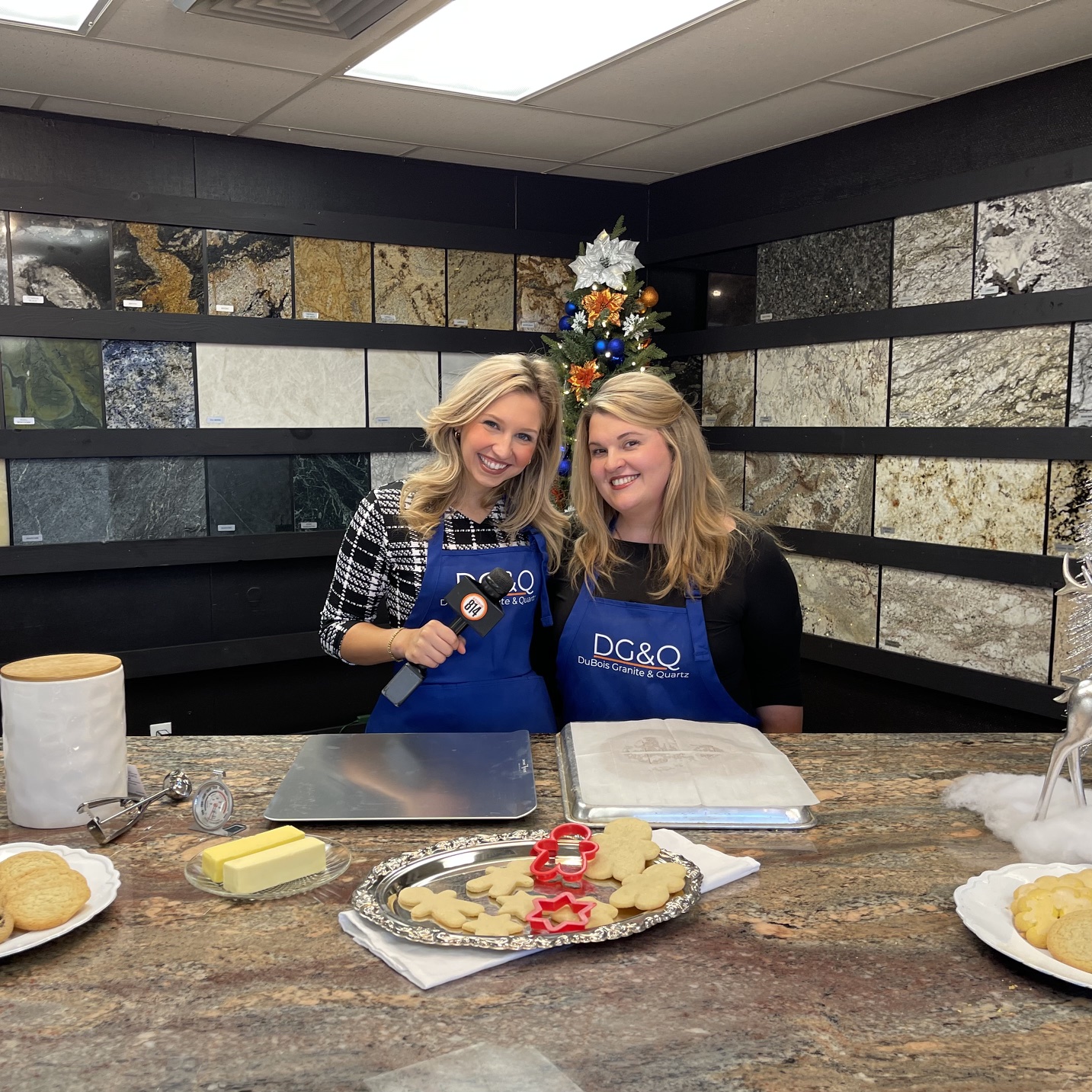 Two women wearing blue aprons stand behind a kitchen counter, smiling, with a decorated cake in front of them.