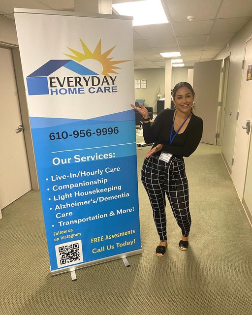 Woman standing next to a large banner for Everyday Home Care, smiling, pointing at the banner, in a hallway.