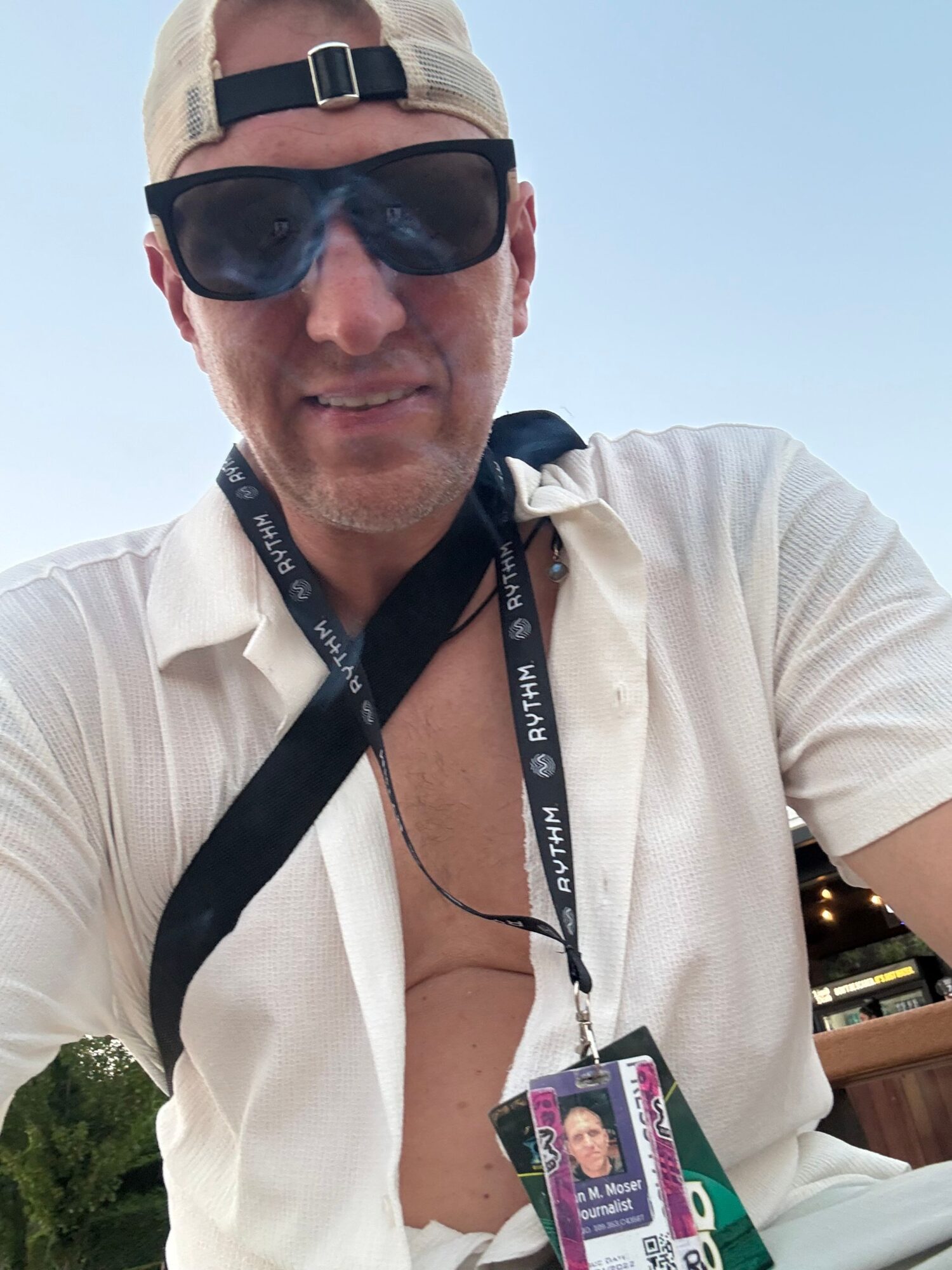 Person wearing sunglasses and a white shirt, sitting outdoors with a lanyard and ID badge, against a blue sky.