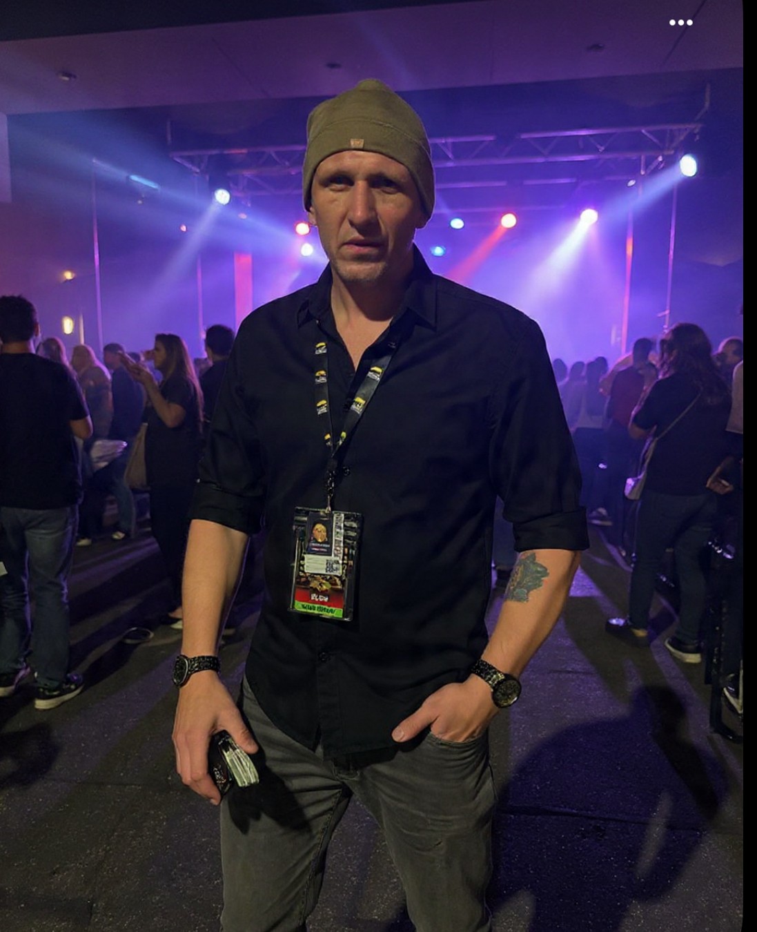 Man in beanie and dark shirt stands in a crowded indoor event with colorful stage lights.