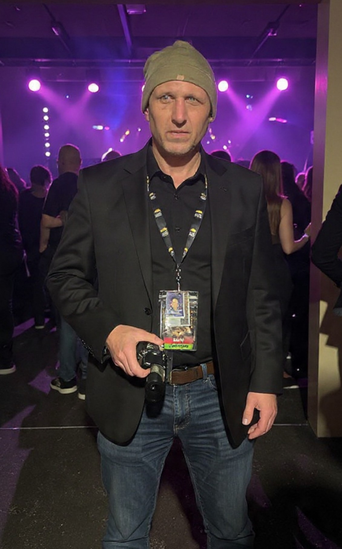 Man in a gray beanie and black blazer holding a microphone at a concert or event, with purple stage lights in background.