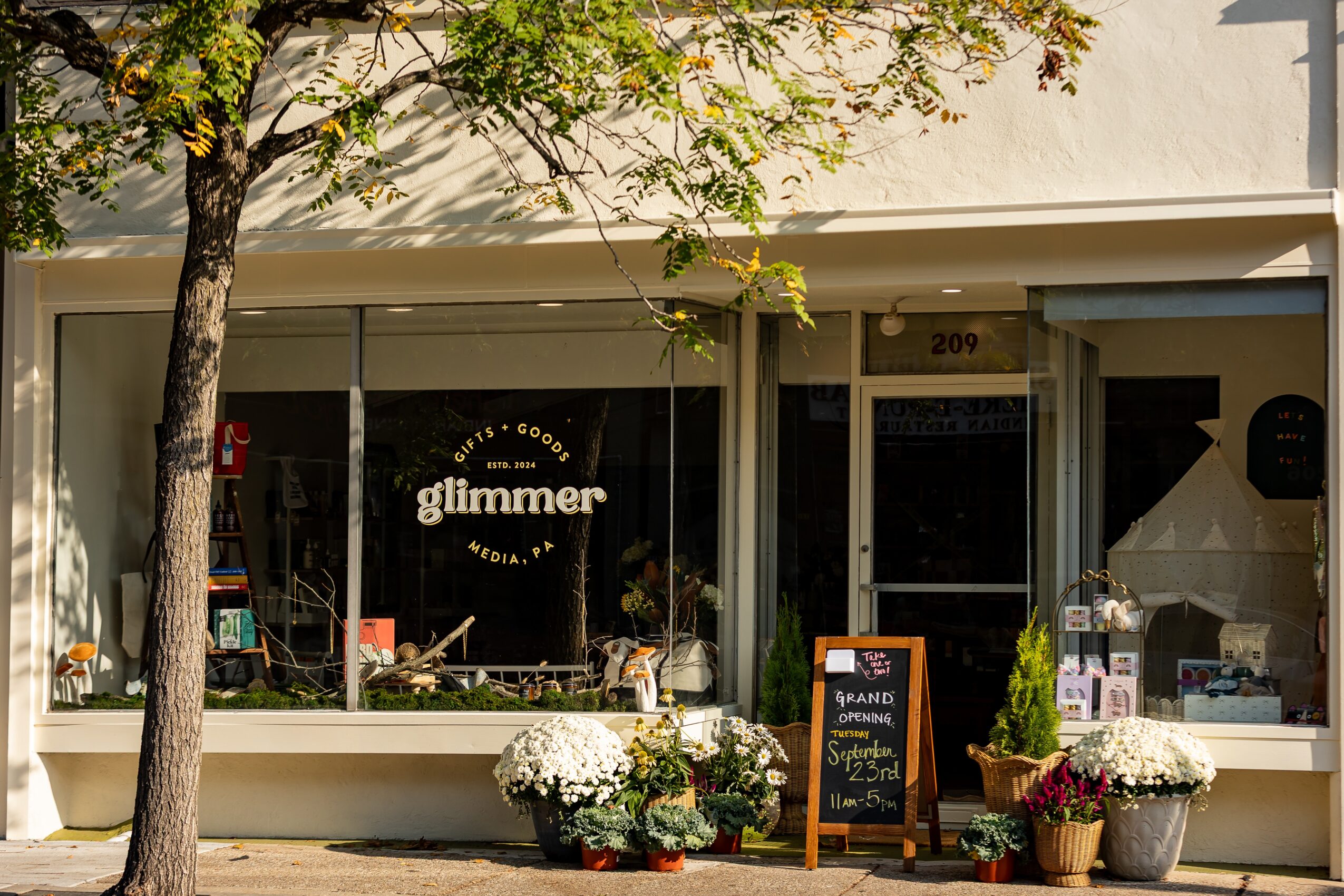 Storefront with a tree in front, displaying the word 'glimmer' on the window, and a chalkboard sign outside.