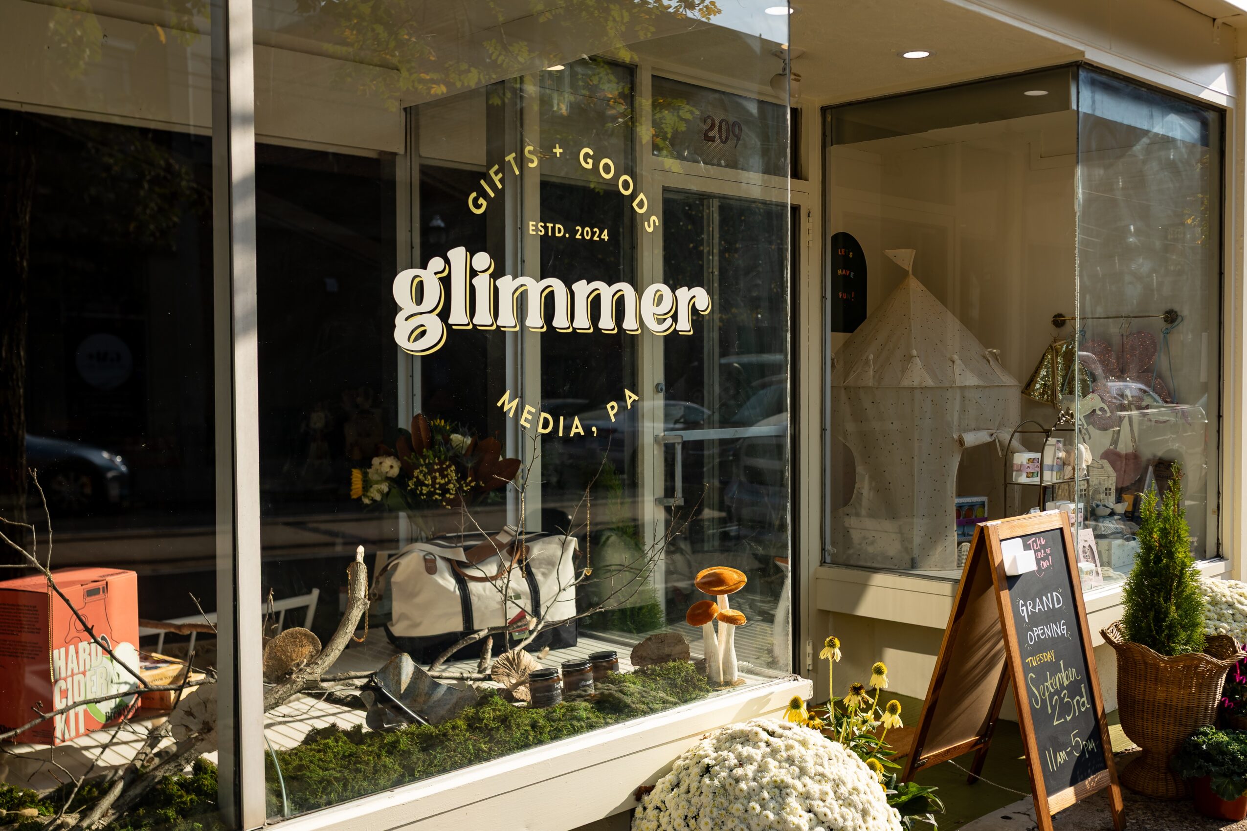 Store window with the word 'glimmer' and signs for gifts, goods, and media, with plants and decorative items inside.