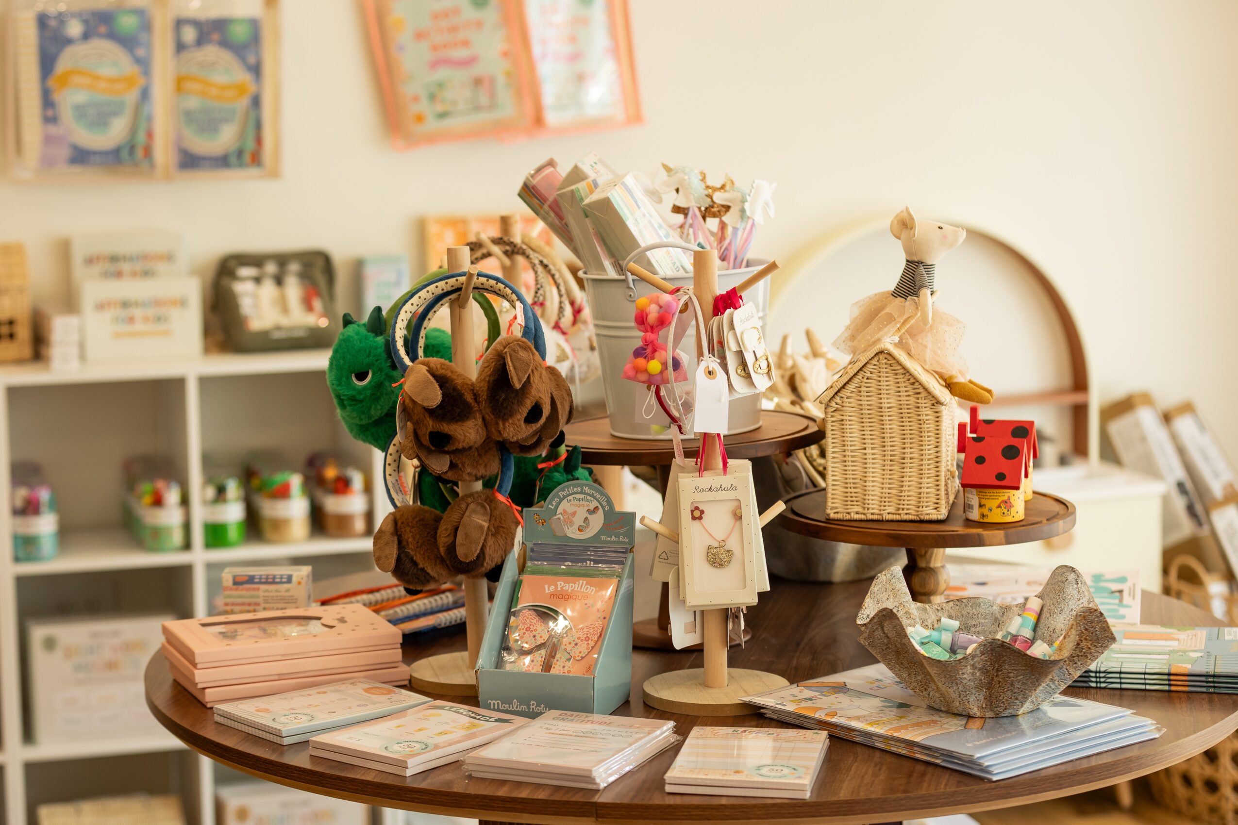 A round table with children's toys, books, and decorative items in a bright room with shelves in the background.