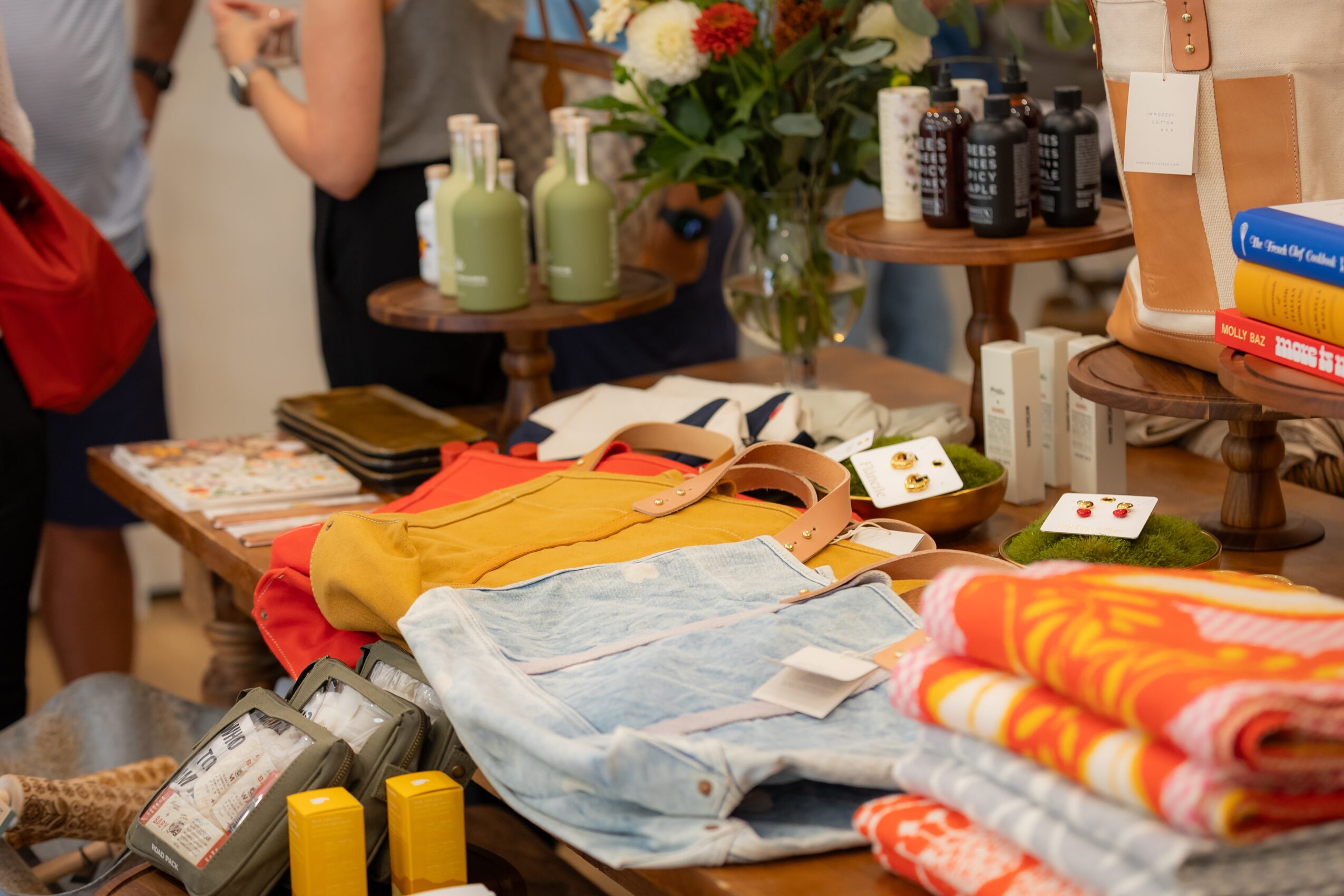 Table with various items including towels, bags, and small bottles, with people and shelves in the background.