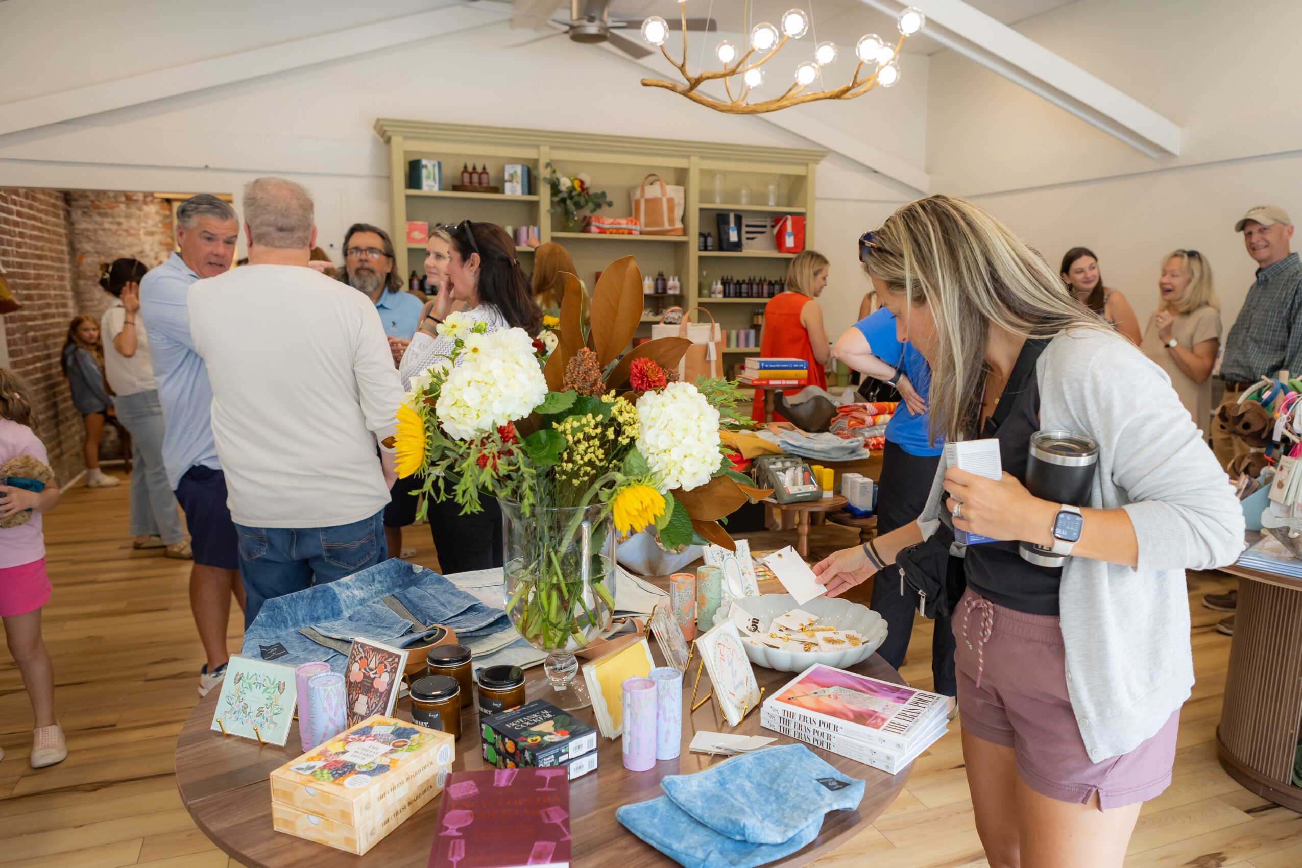 People gather around a table with flowers and various items in a well-lit room with shelves and a chandelier.