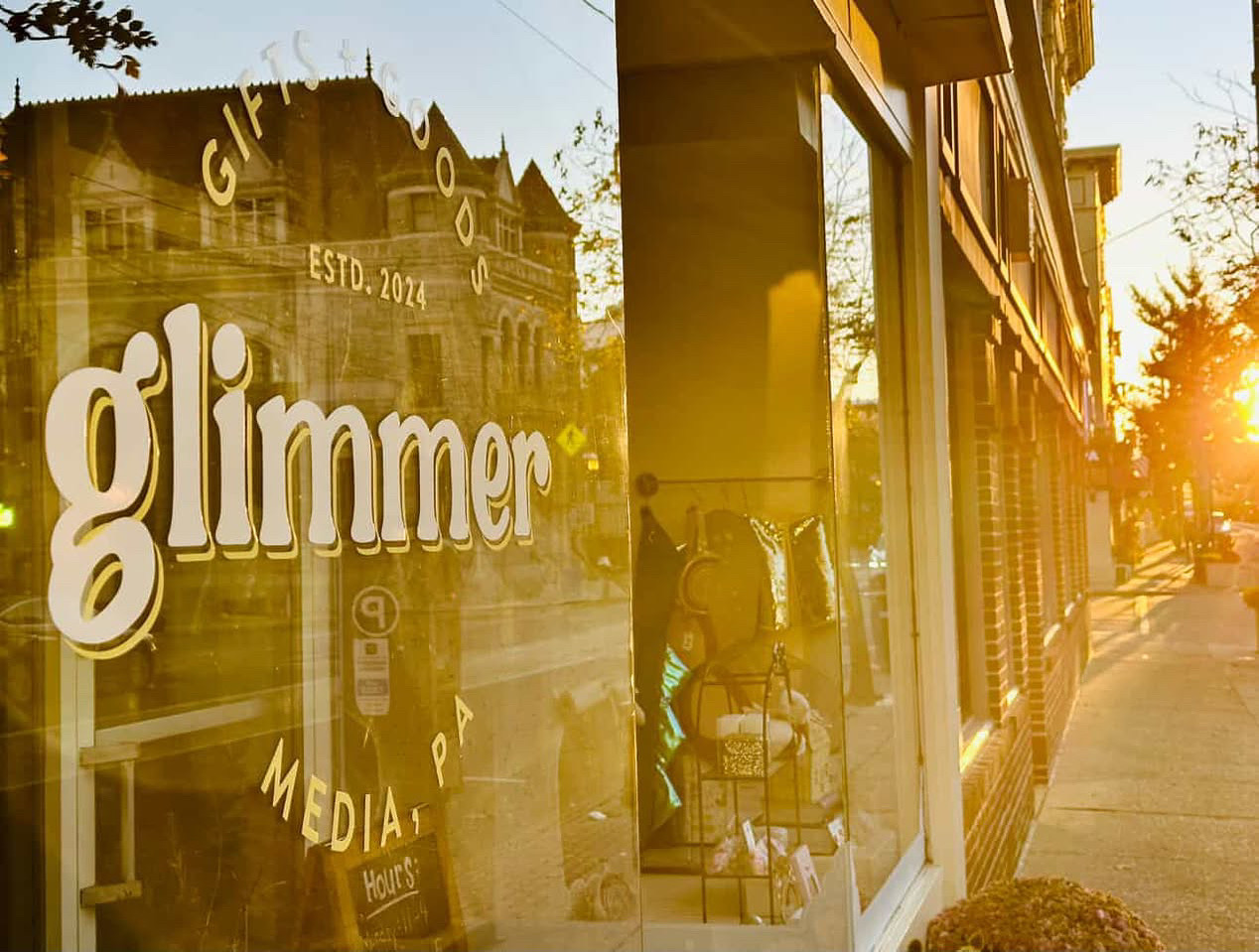 Store window reflecting street scene with sunlight, buildings, and a person inside, with the word 'glimmer' on the glass.