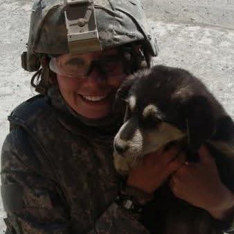Person in military gear smiling and holding a black and white puppy outdoors.