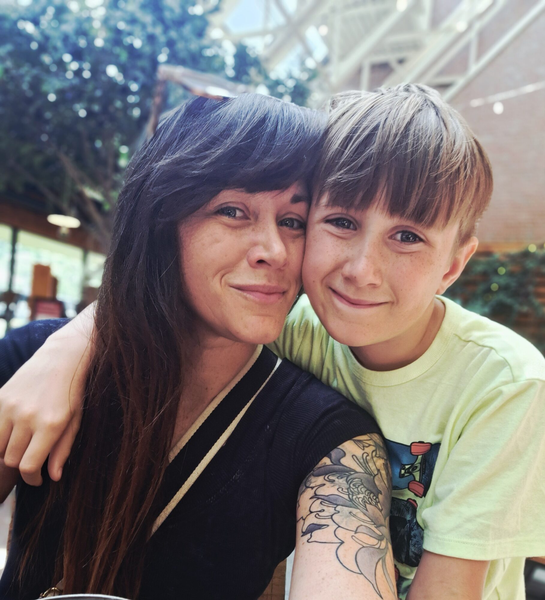 Woman and boy smiling, close together, outdoors with trees and structure in background.