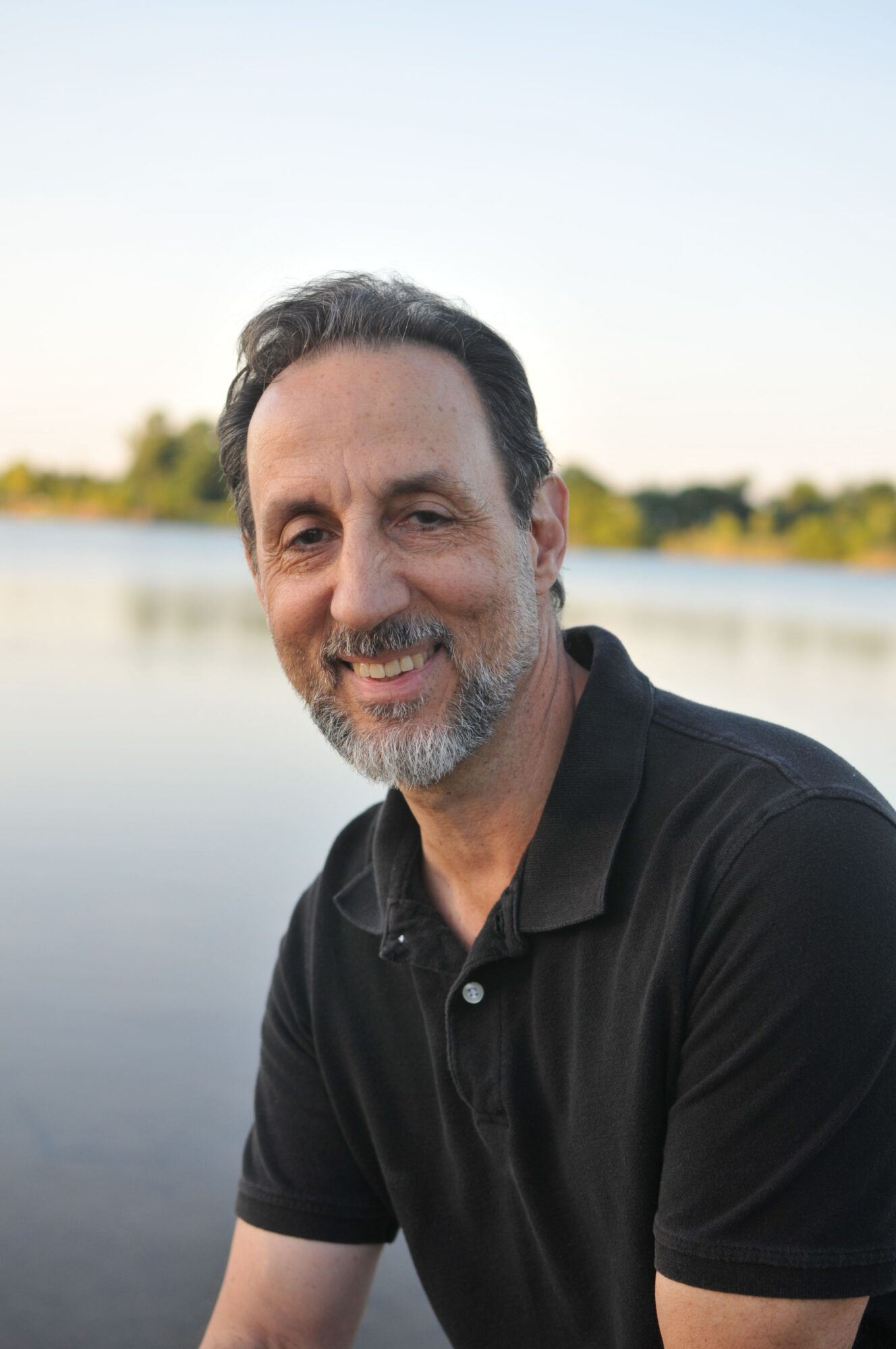 Man with dark hair and beard wearing a black shirt outdoors near water, smiling slightly.