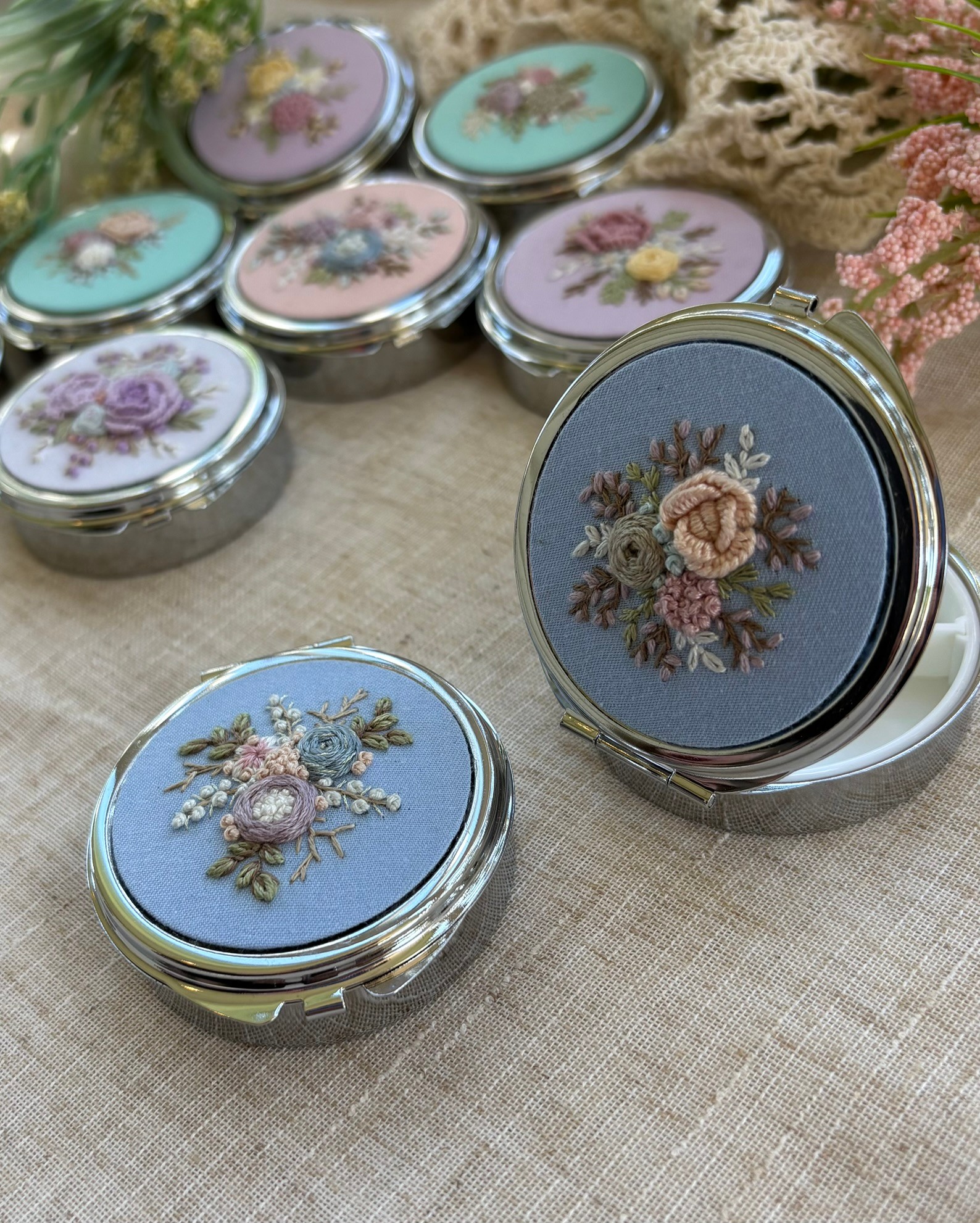 Round compact mirrors with floral embroidery on pastel blue and pink backgrounds, arranged on a fabric surface with flowers nearby.